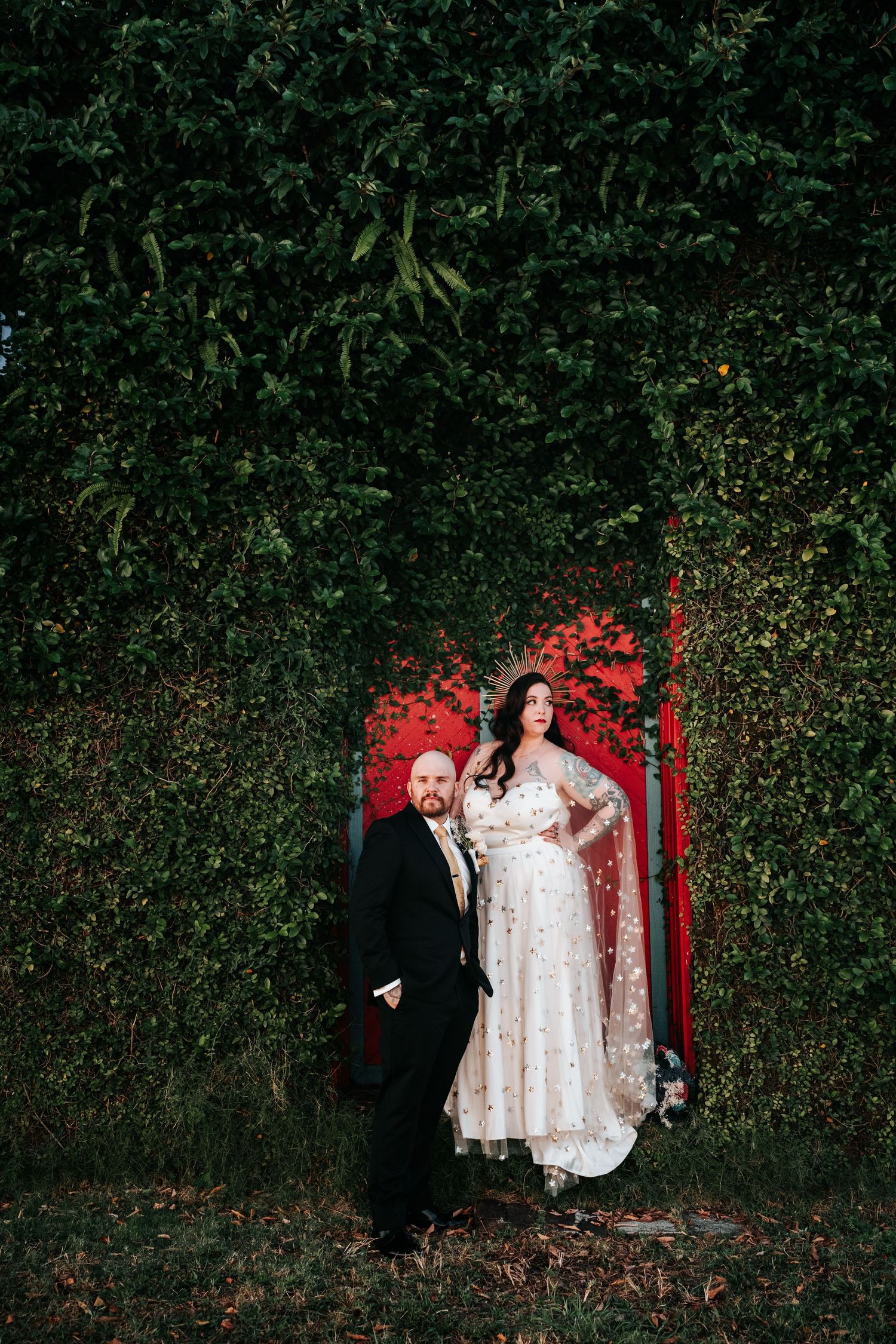 A couple stands in front of a lush green hedge wall with a vibrant red door frame creating a striking contrast.