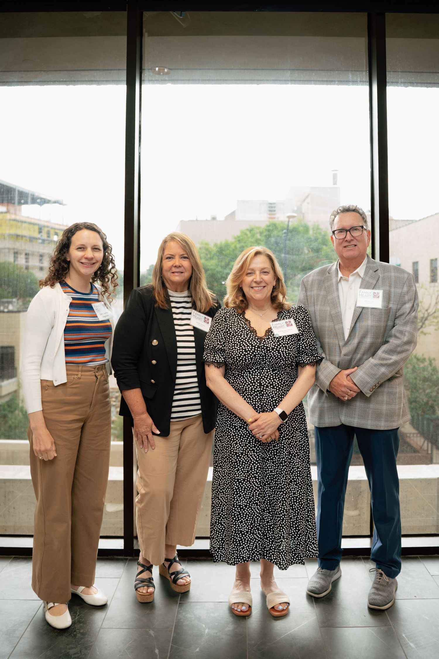 Four professionals smiling together in front of large windows at a corporate event, wearing name badges.