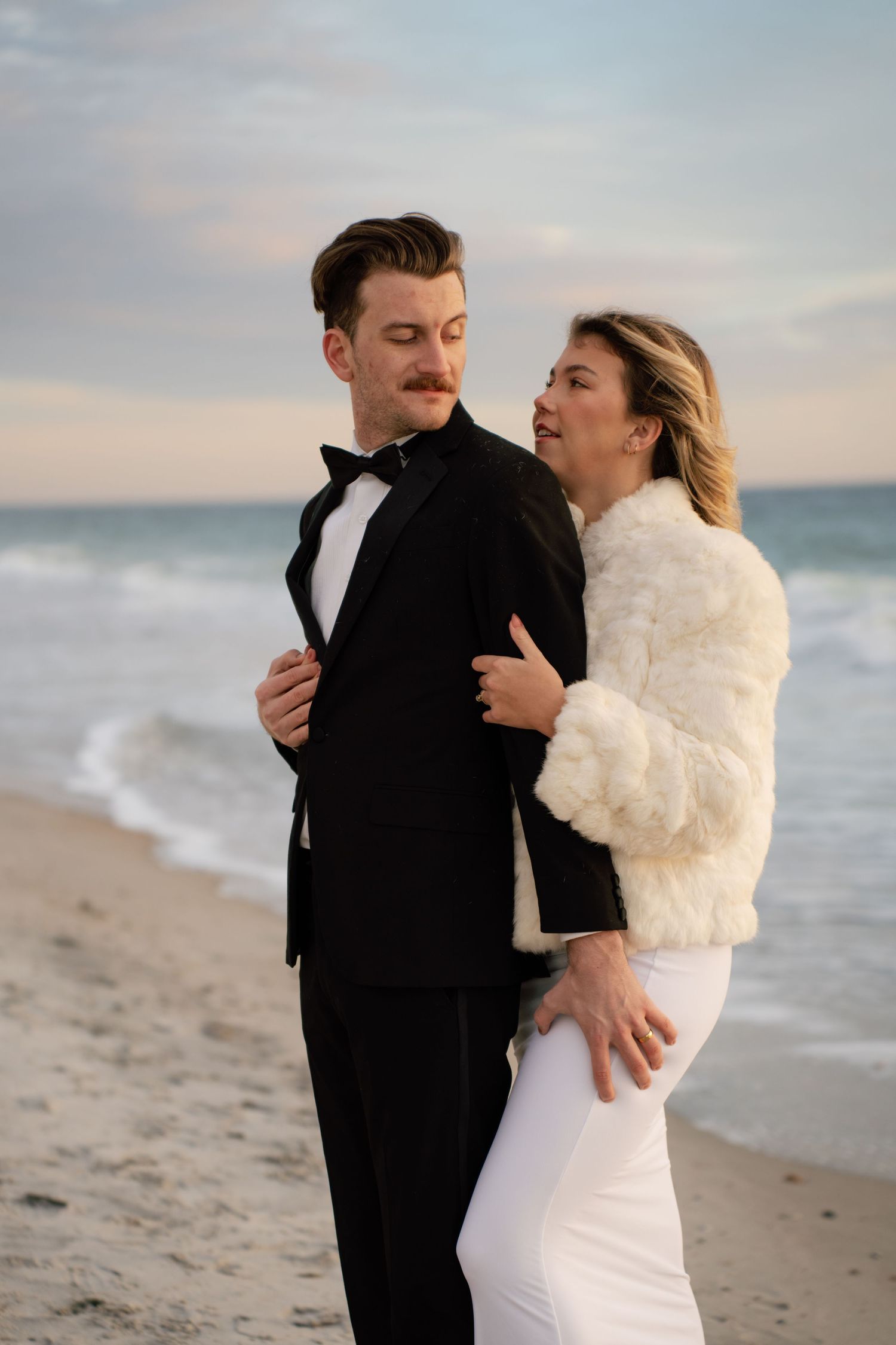 Couple in formal wedding attire embrace and laugh together on a beach at sunset.