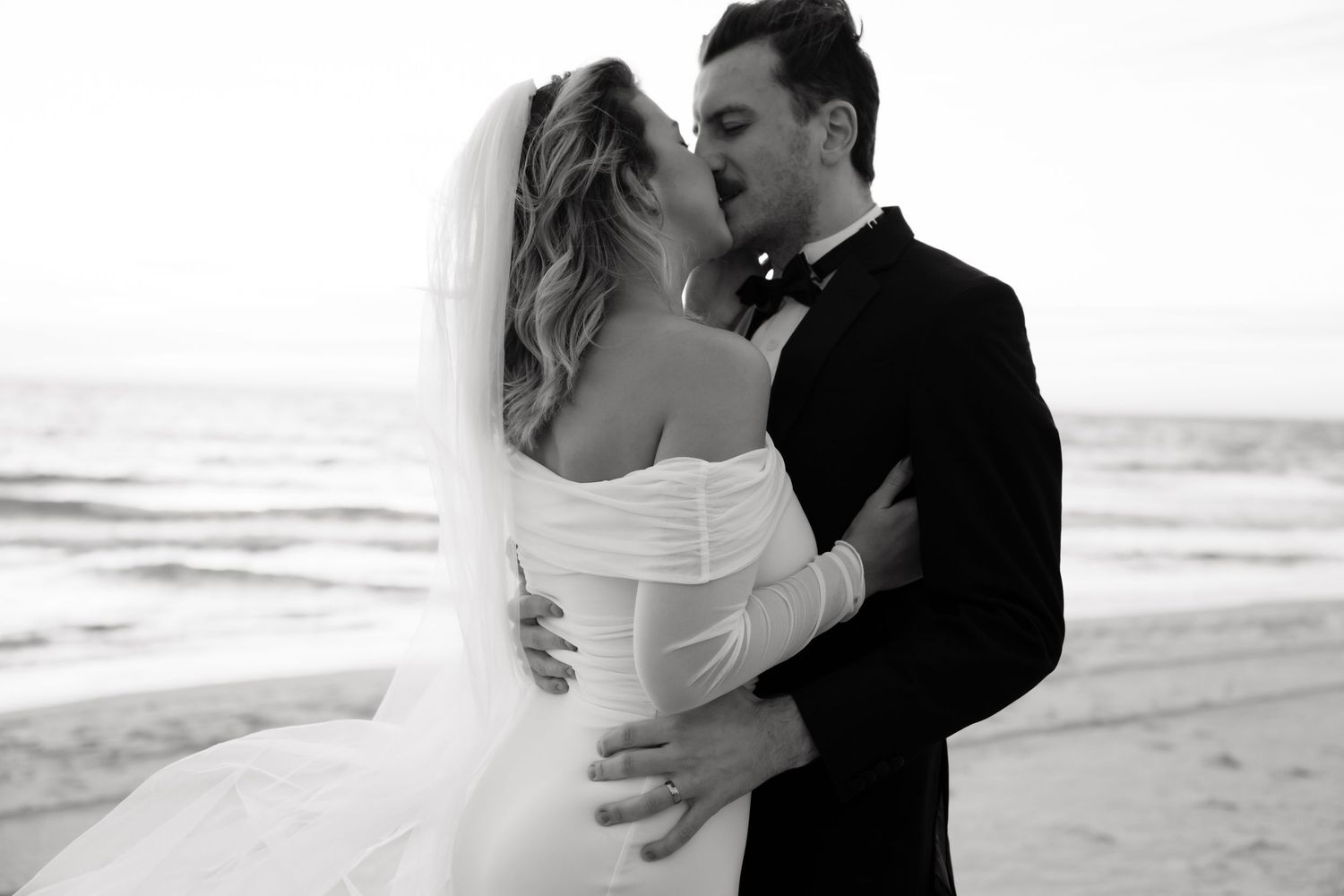 A couple in formal wedding attire shares a romantic kiss on the beach at sunset.