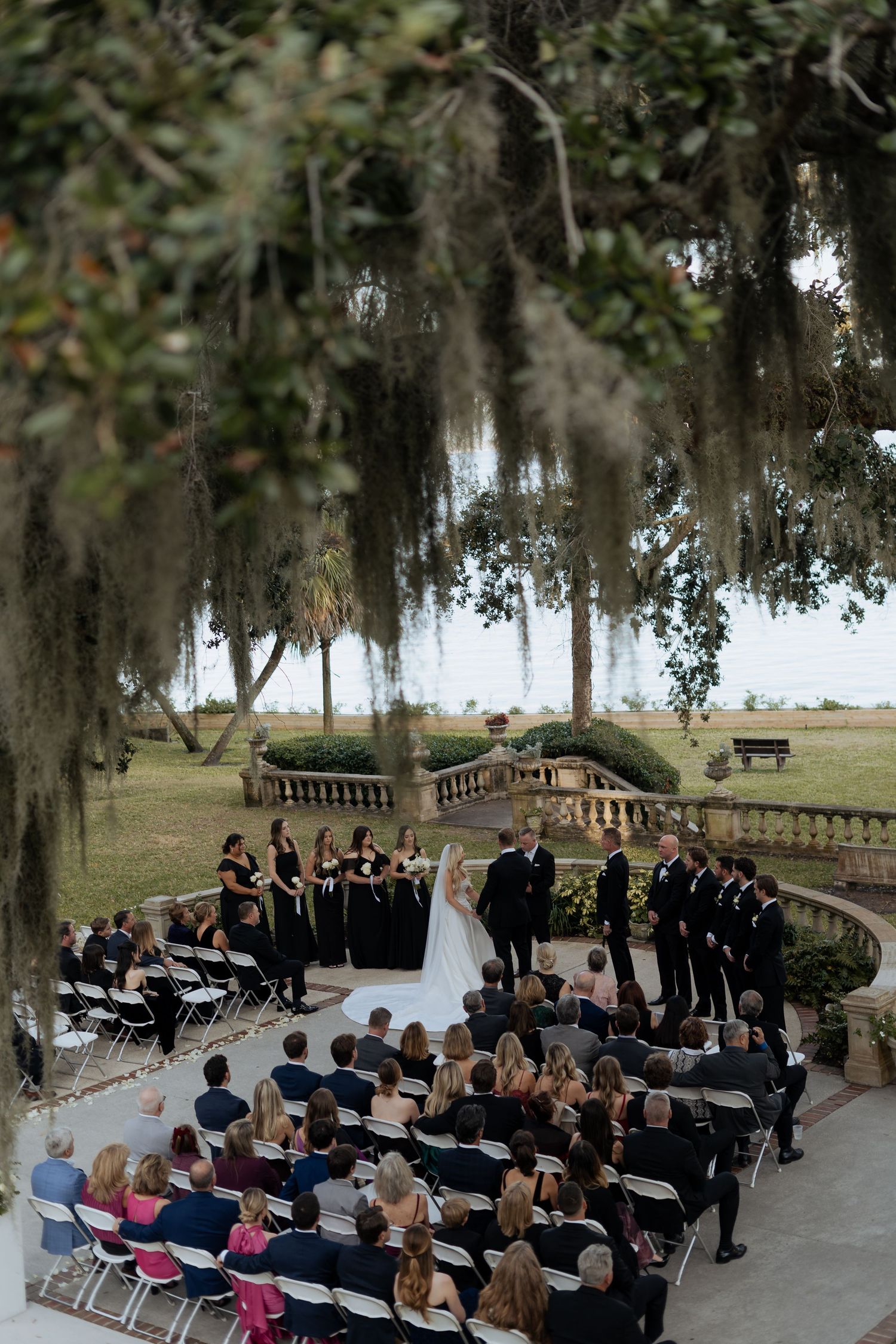 Outdoor wedding ceremony setup under moss-draped oak trees with rows of white chairs facing a waterfront view.