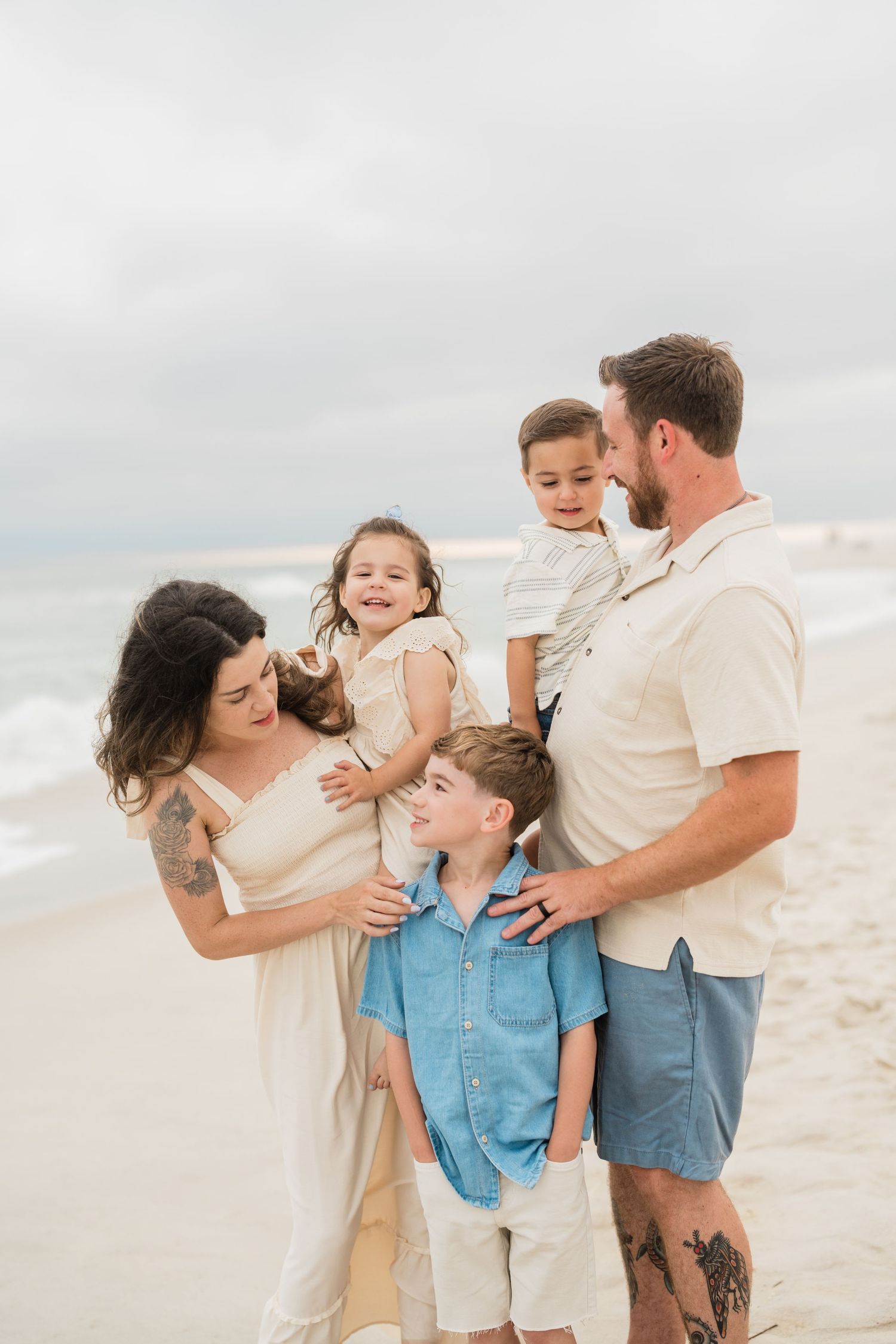Family of five smiling together on a beach, parents holding children, dressed in neutral tones on a cloudy day.