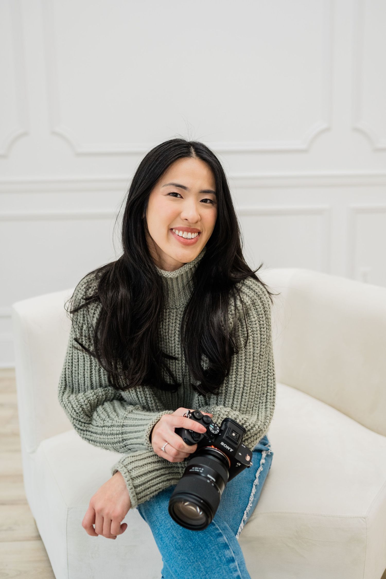 Photographer in grey sweater sitting on white couch with camera in casual studio setting.