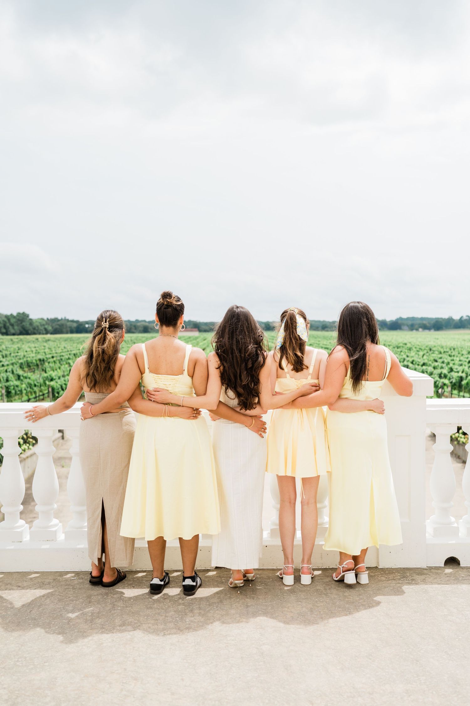 Bridesmaids in cream dresses standing arm in arm overlooking scenic landscape.