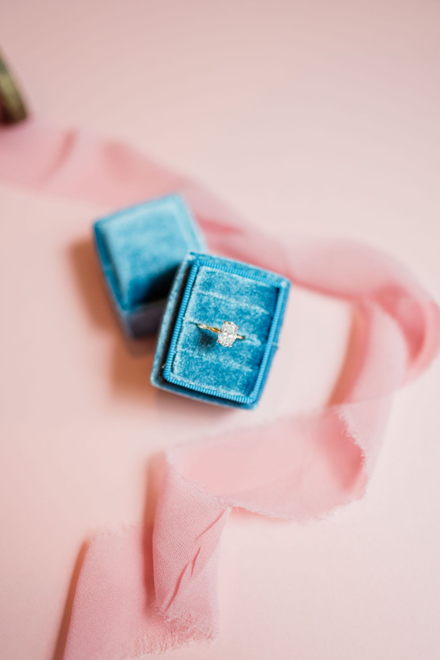Blue velvet ring box with pink ribbon on soft pink background.