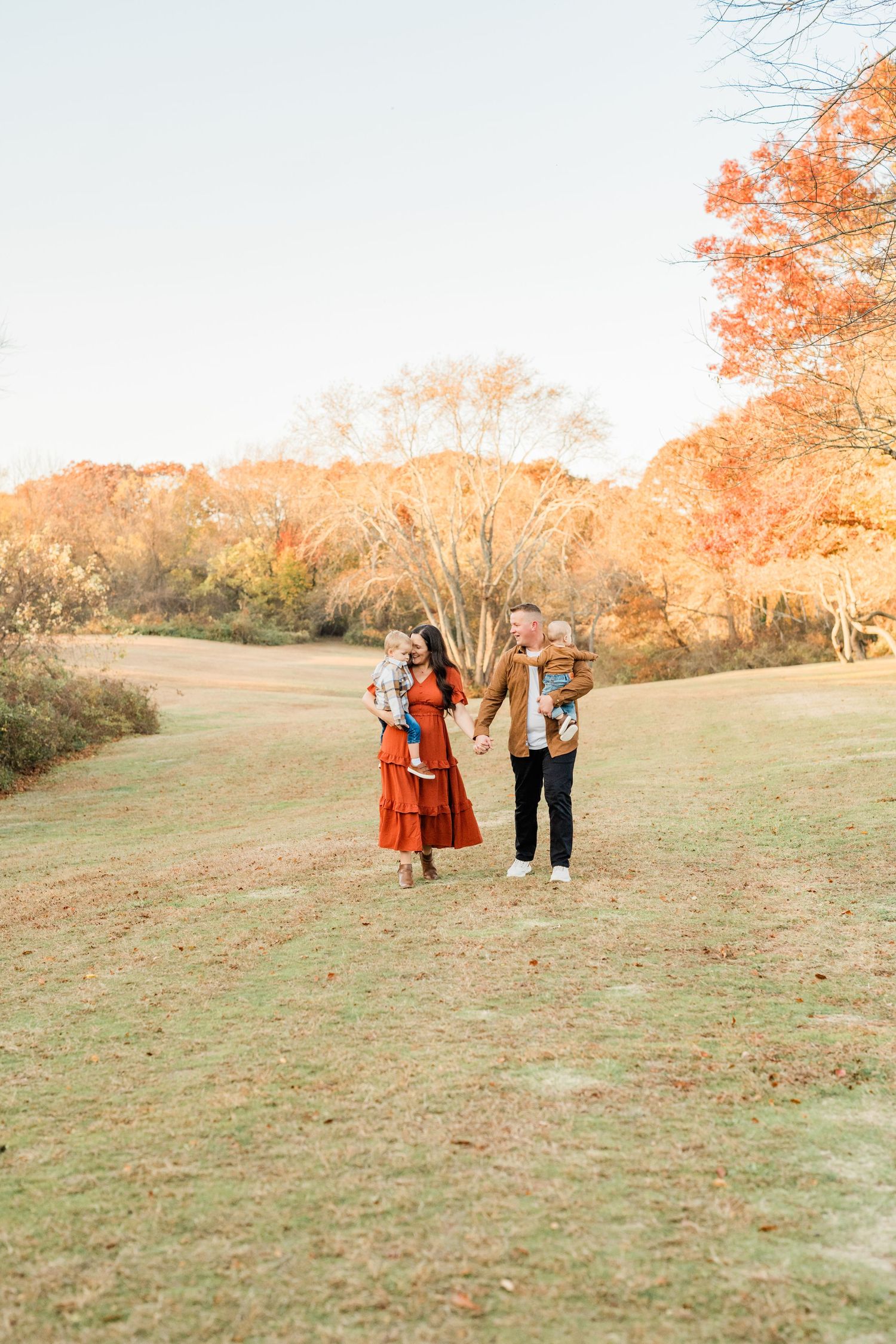 Family of four walking hand-in-hand through a lush green field surrounded by vibrant autumn foliage.