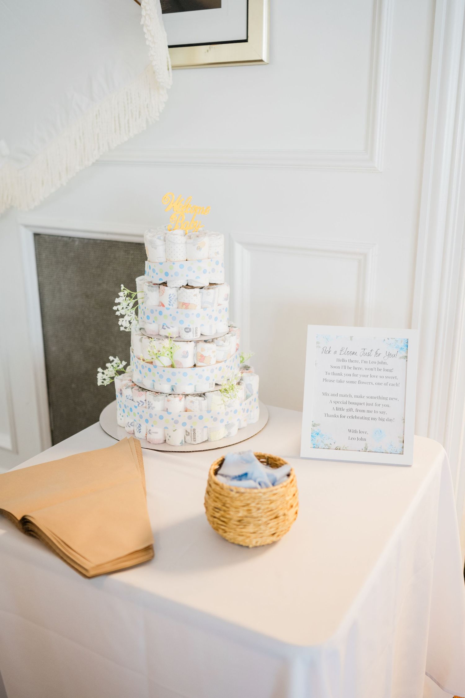 White and blue diaper cake display with floral decorations and guest book on white table.