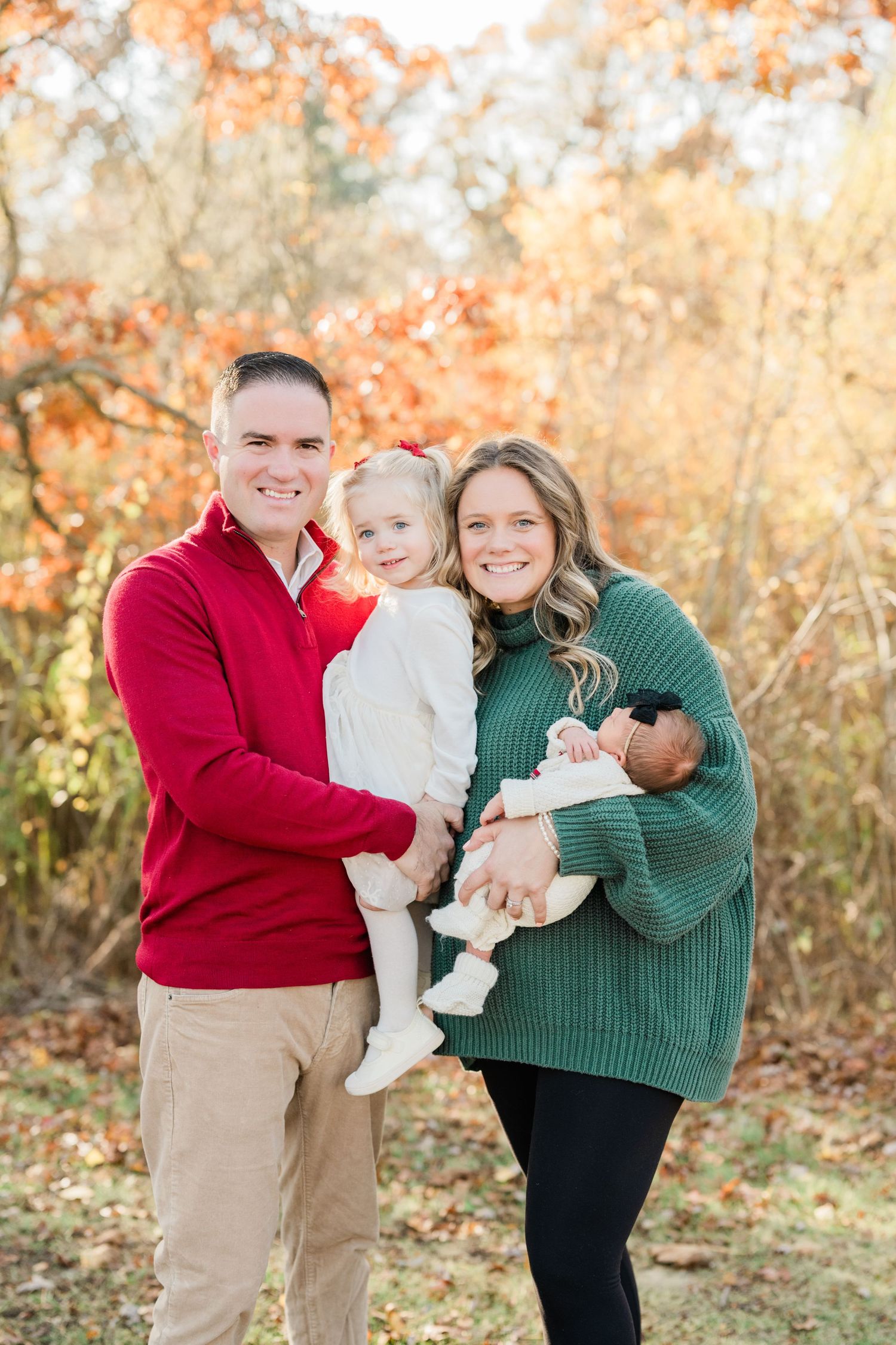 Family of four posing outdoors in fall foliage, parents holding toddler girl and newborn baby, wearing red and green.