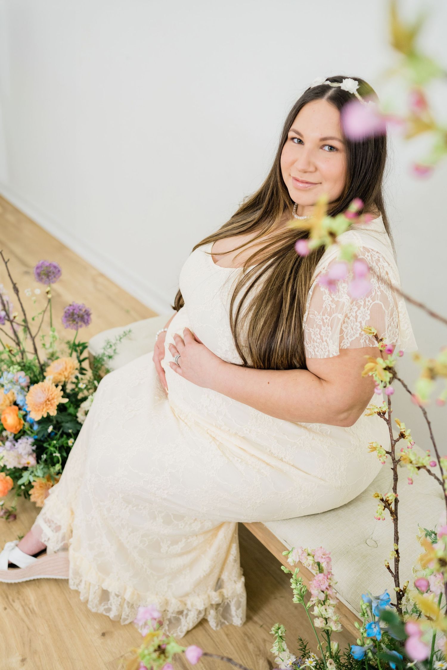 Elegant maternity session with lace dress and colorful spring flowers in studio setting.