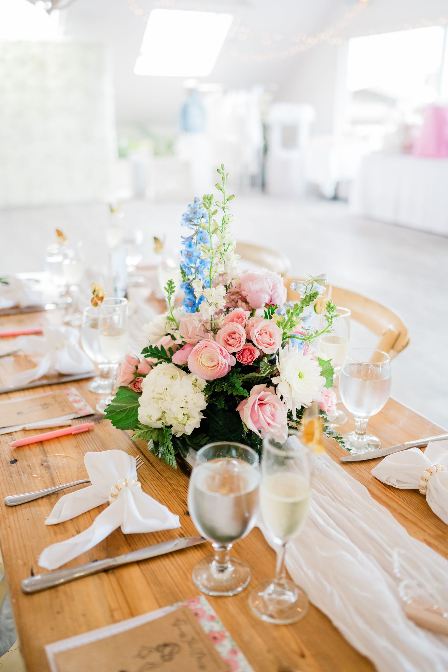 Table setting with pink and blue floral centerpiece, wine glasses, and white linens on wooden surface.