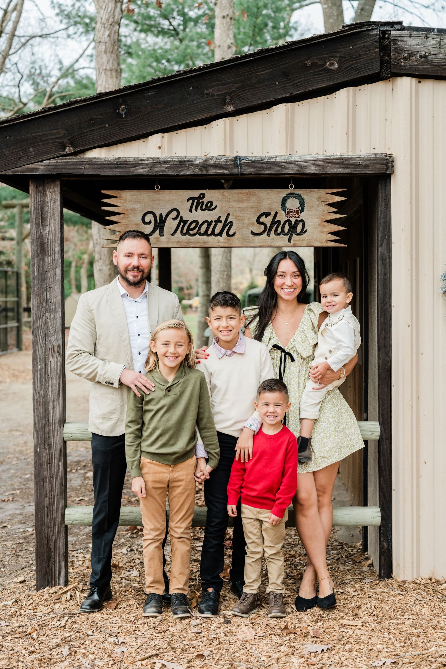 Family portrait outside The Wreath Shop with rustic wooden entrance and pine trees in background.