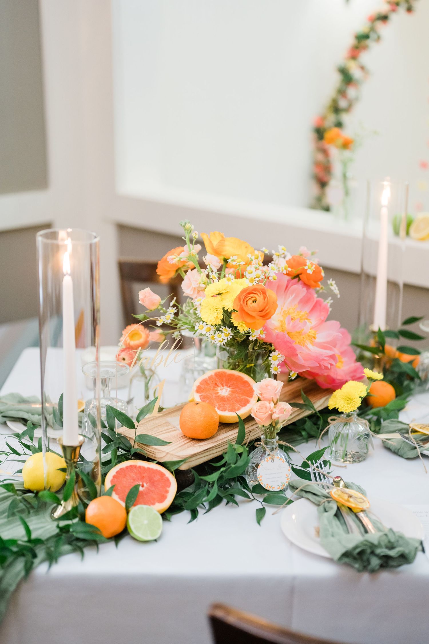 Elegant table centerpiece with citrus fruits, flowers, and candles in peach and coral tones.