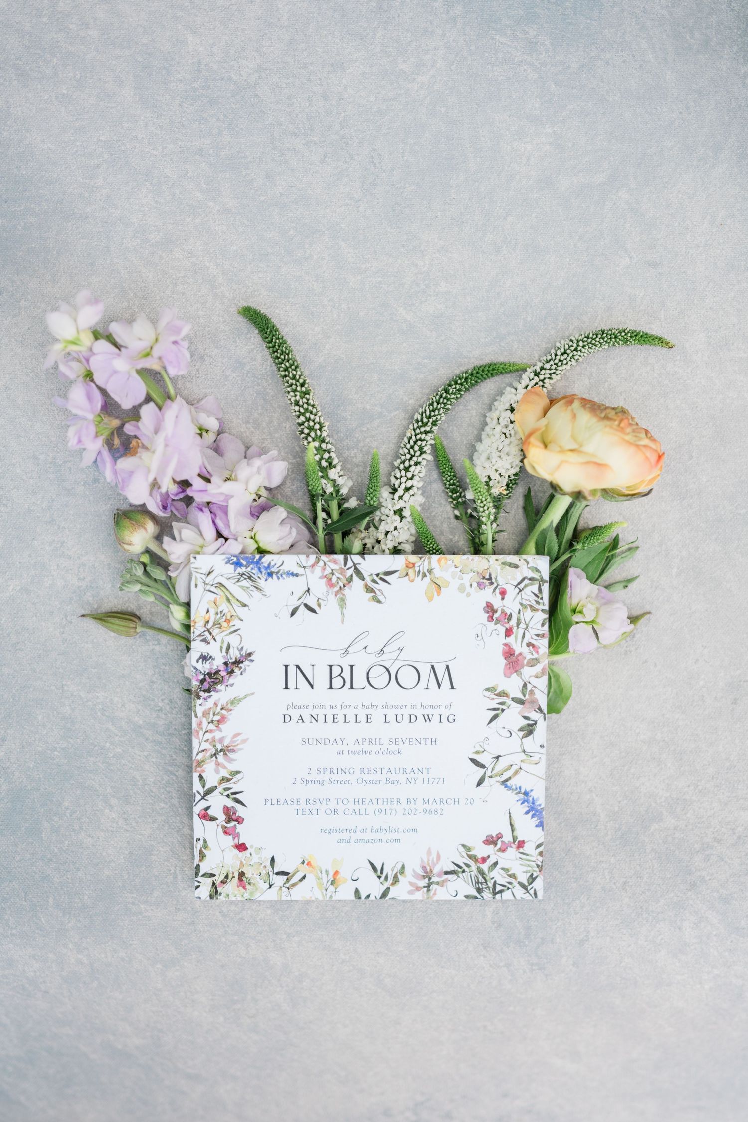 Floral wedding invitation card with 'In Bloom' text surrounded by fresh flowers and greenery on grey background.