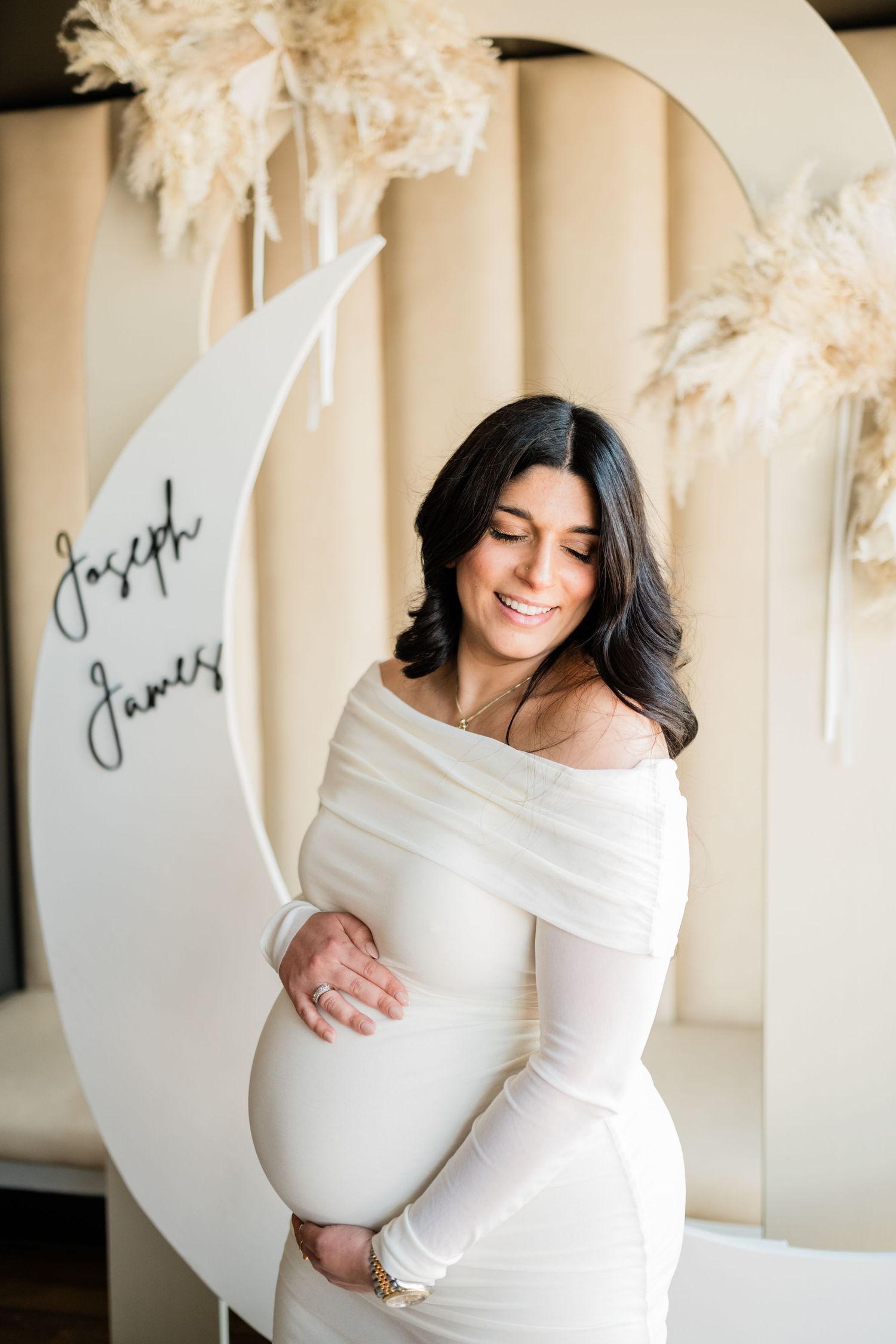 Maternity photo of woman in white dress posing against curved backdrop with text.