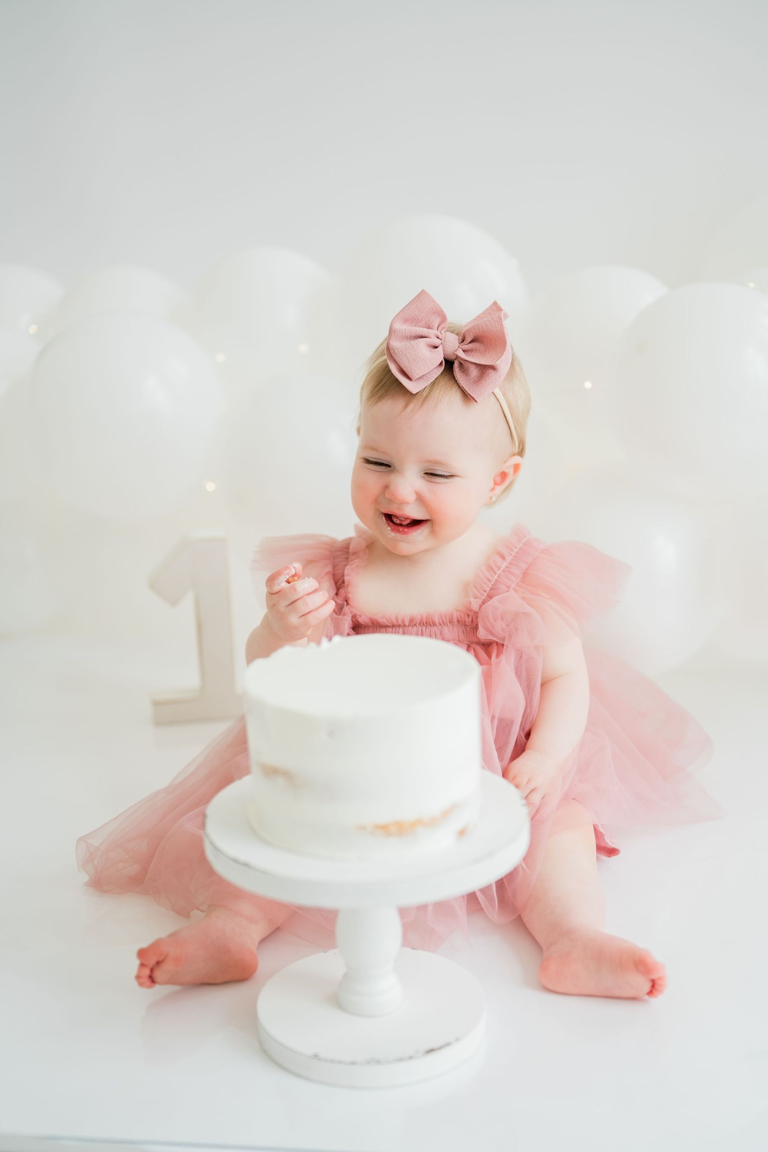 Baby girl in pink tutu smashes her first birthday cake, wearing a bow headband with white balloons in background.