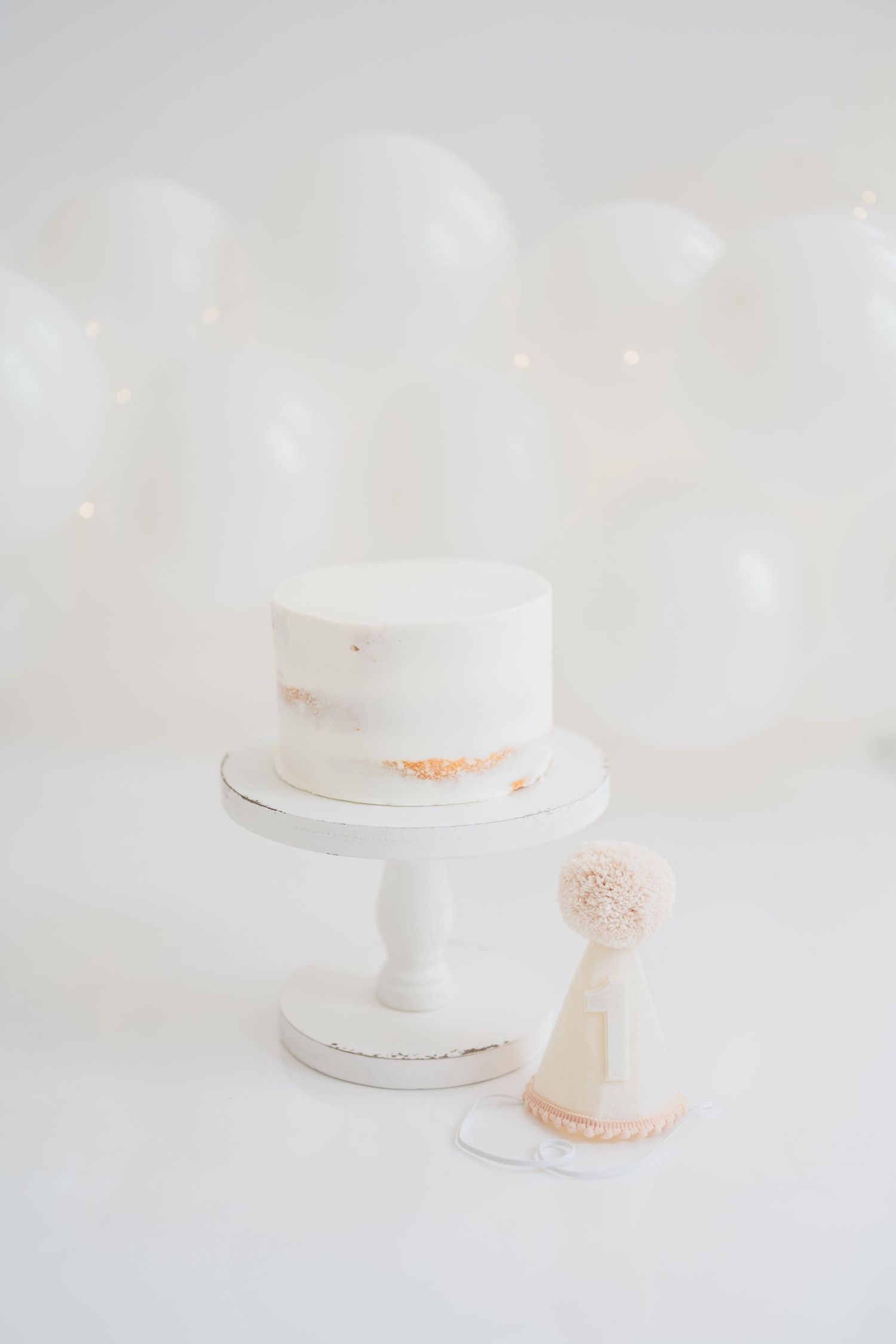 White birthday cake on pedestal stand with party hat and balloons, minimalist first birthday smash cake setup.