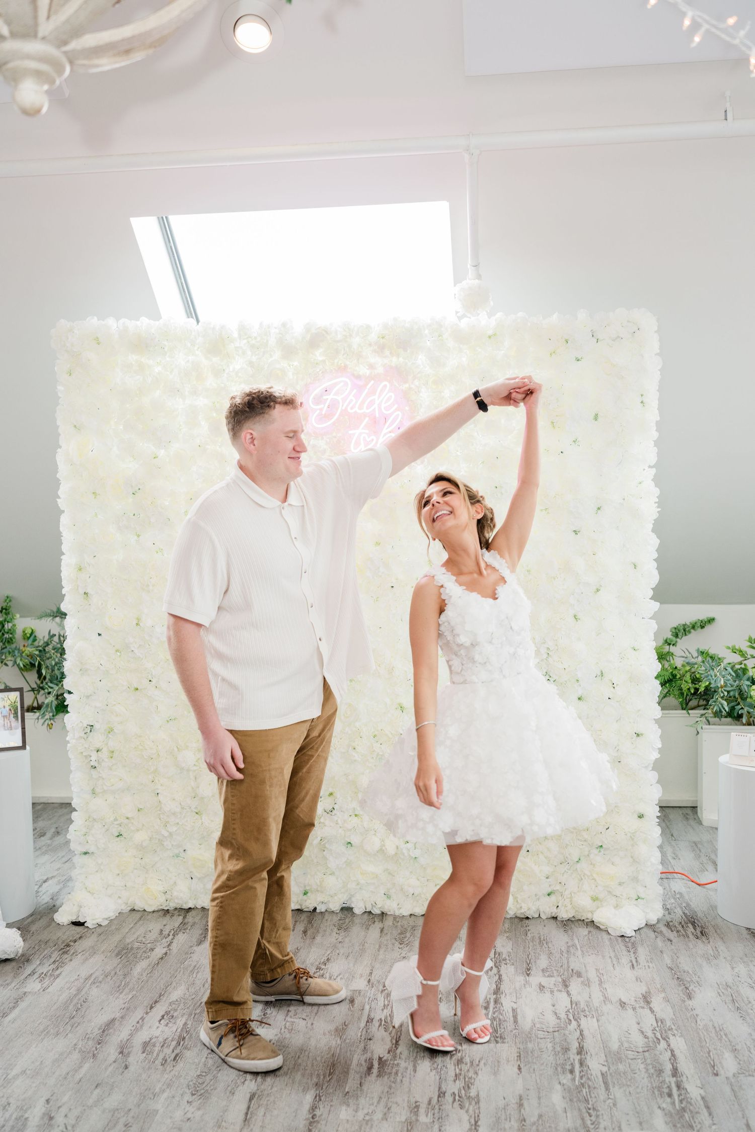 A couple dances playfully in front of a white floral backdrop in a bright studio setting.