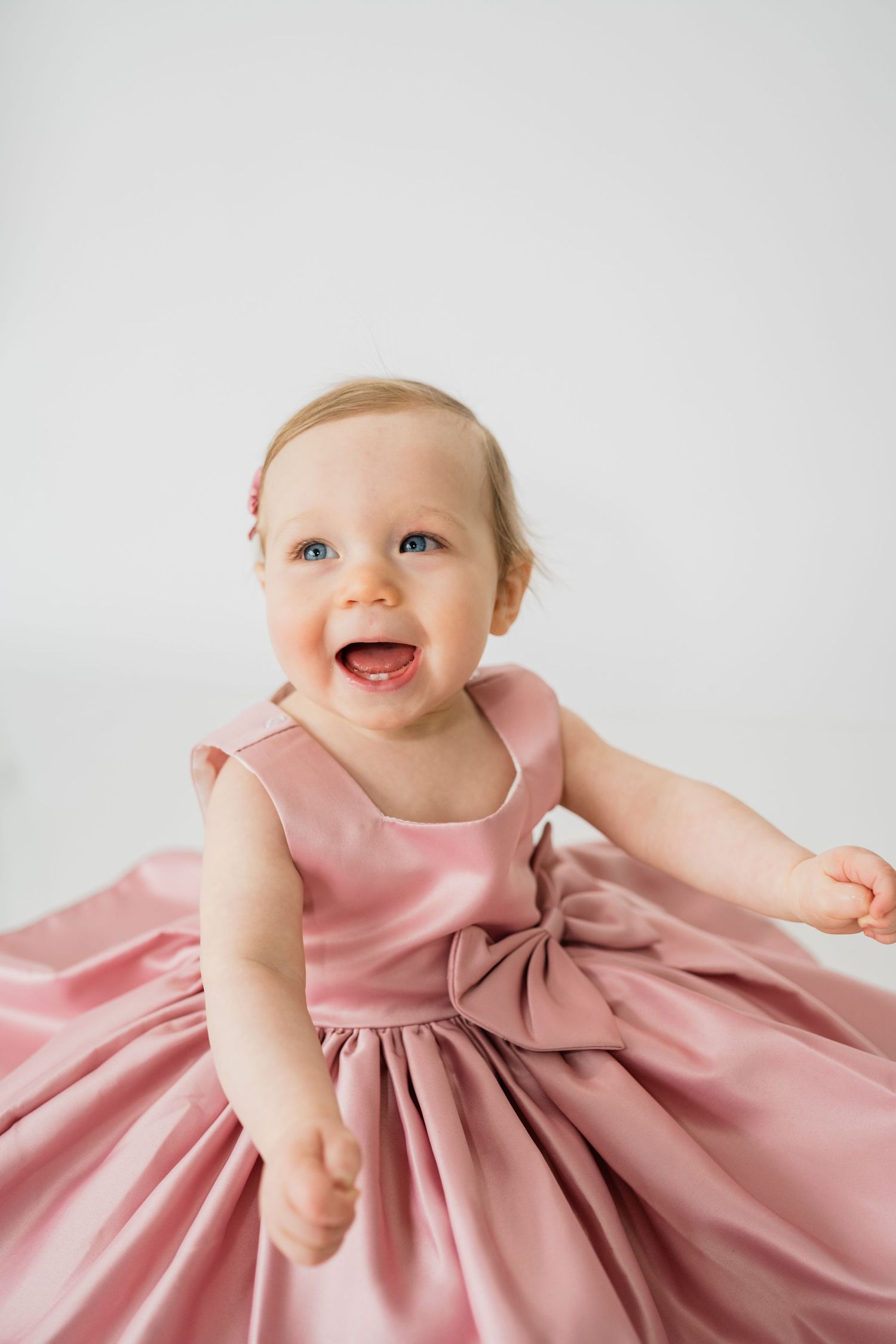 A baby in a pink dress smiles joyfully during a portrait session against a white background.