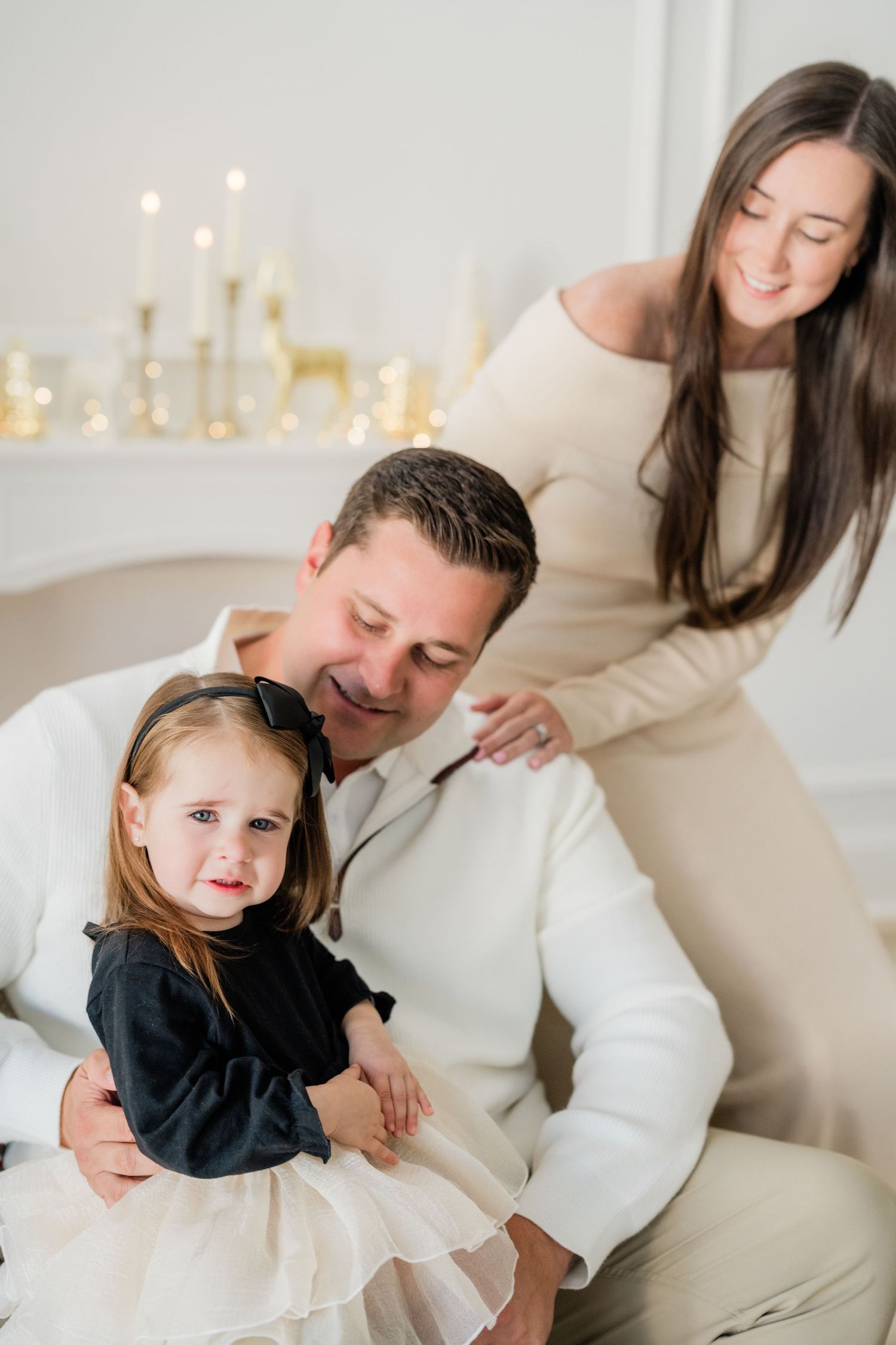 A happy family portrait with a man, woman, and toddler girl posing near a candlelit fireplace in elegant attire.