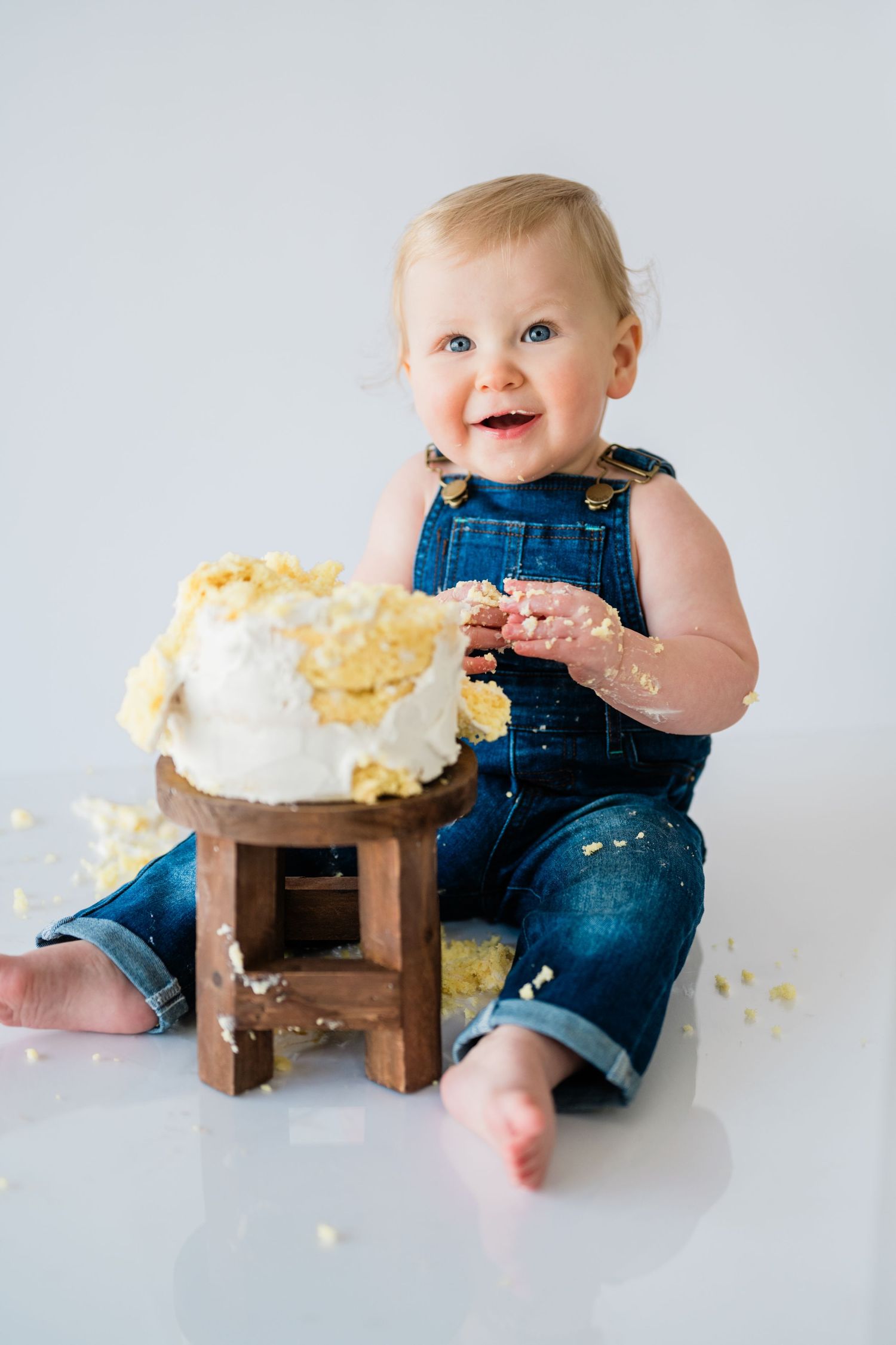 A messy cake smash photo session featuring a baby in denim overalls enjoying white frosting.