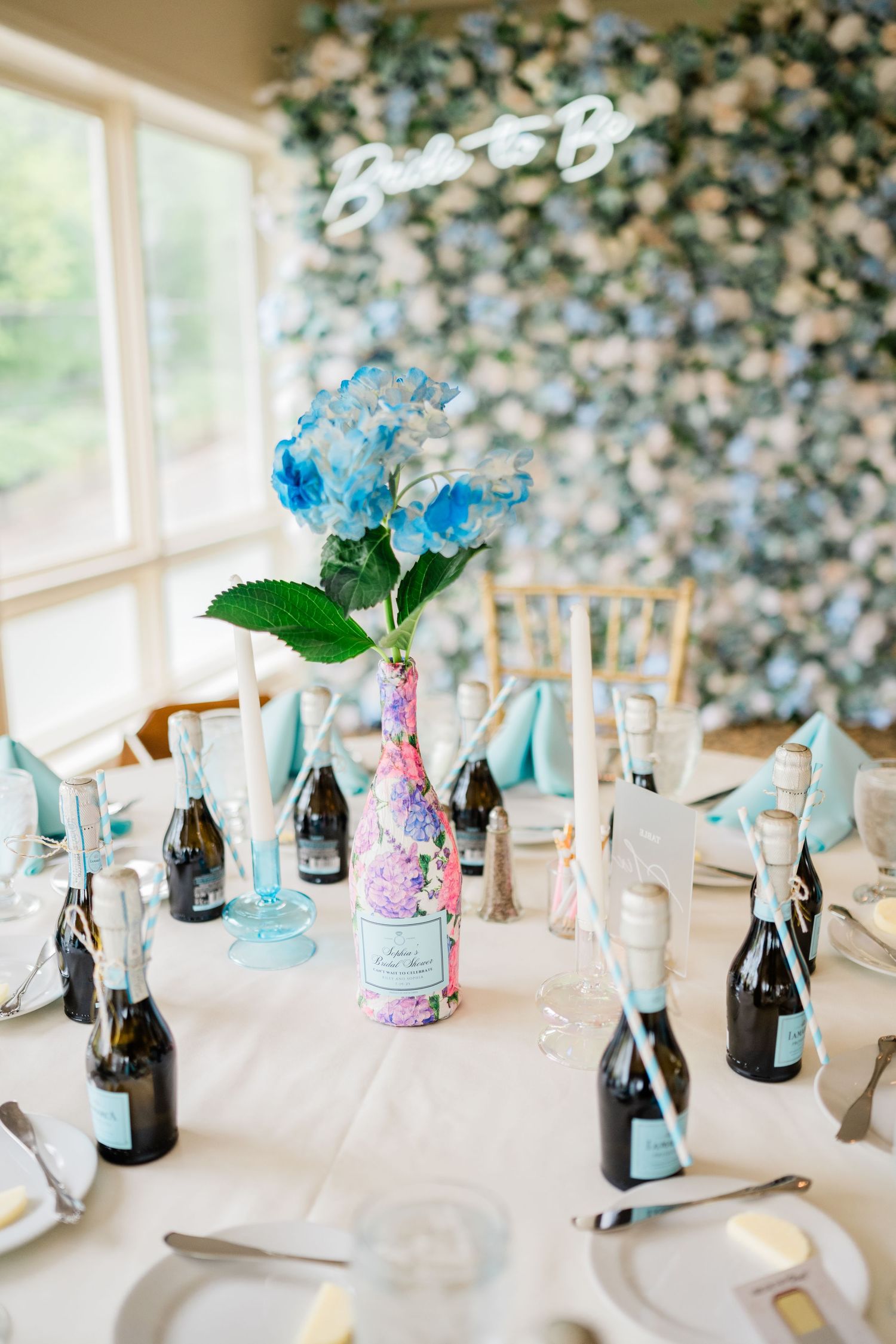 Wedding table decorated with blue hydrangeas in glass vases and mini champagne bottles.