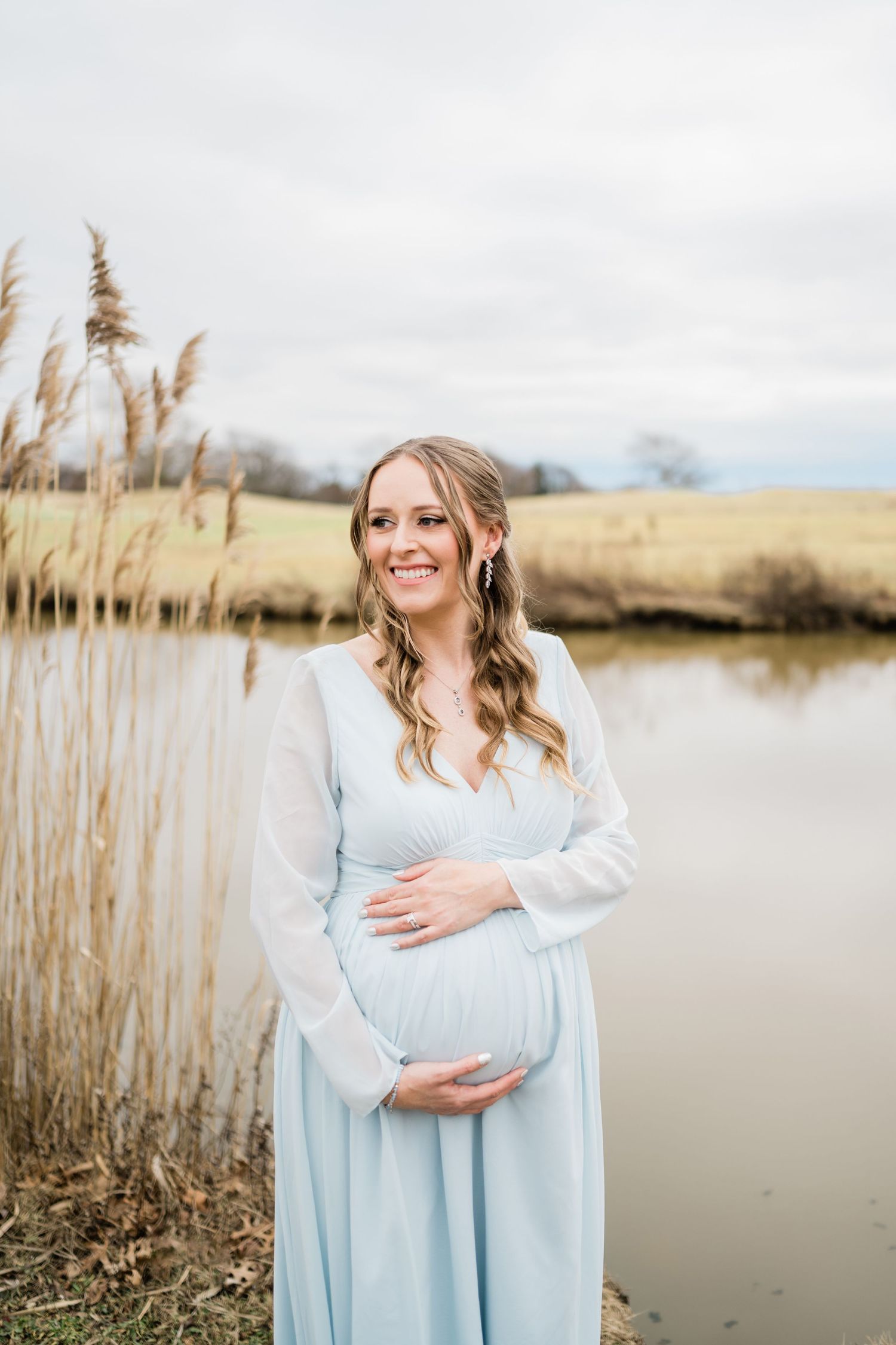 A person in a light blue dress poses by a pond with cattails during a maternity photoshoot.