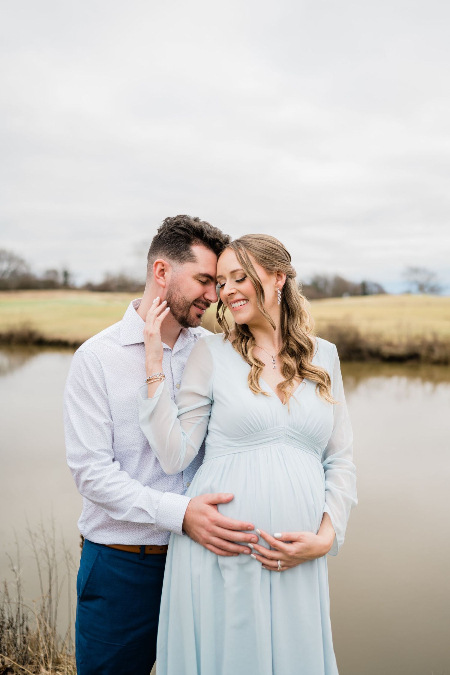 Maternity couple poses by waterfront in soft blue dress during cloudy day.