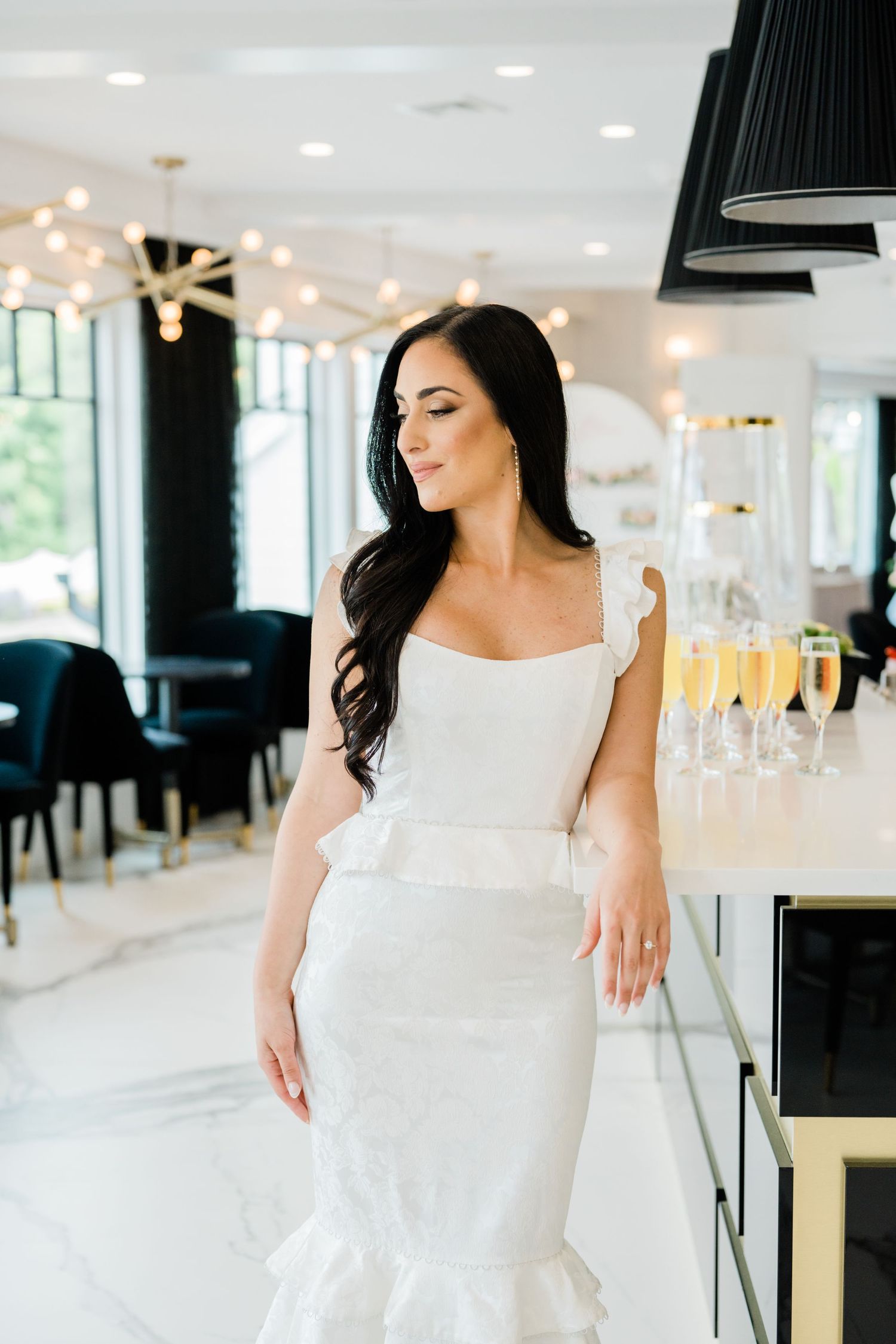 A woman in an elegant white dress poses in a modern restaurant setting with champagne glasses in the background.