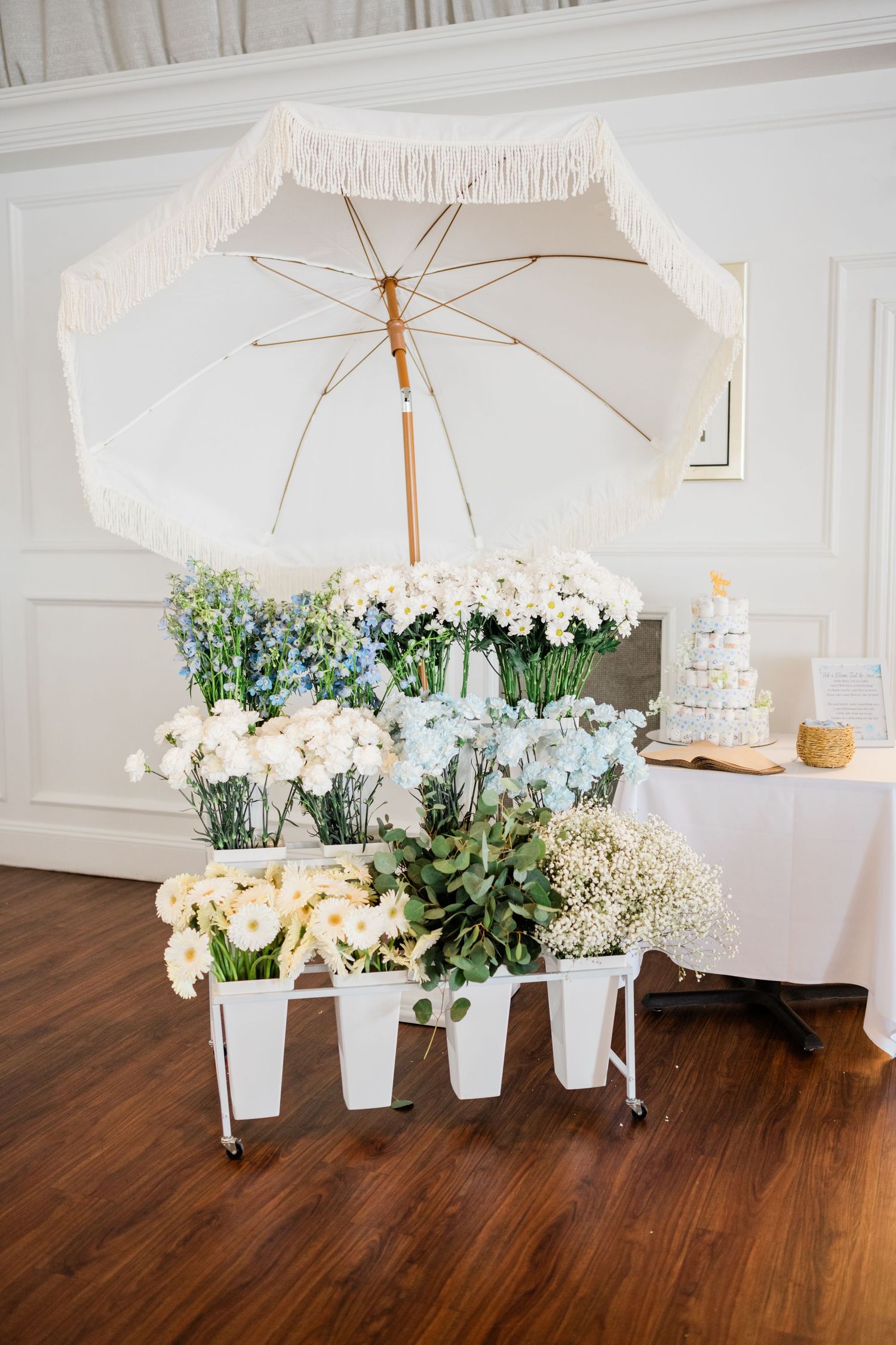 White parasol display with floral arrangements in white containers on wooden floor.