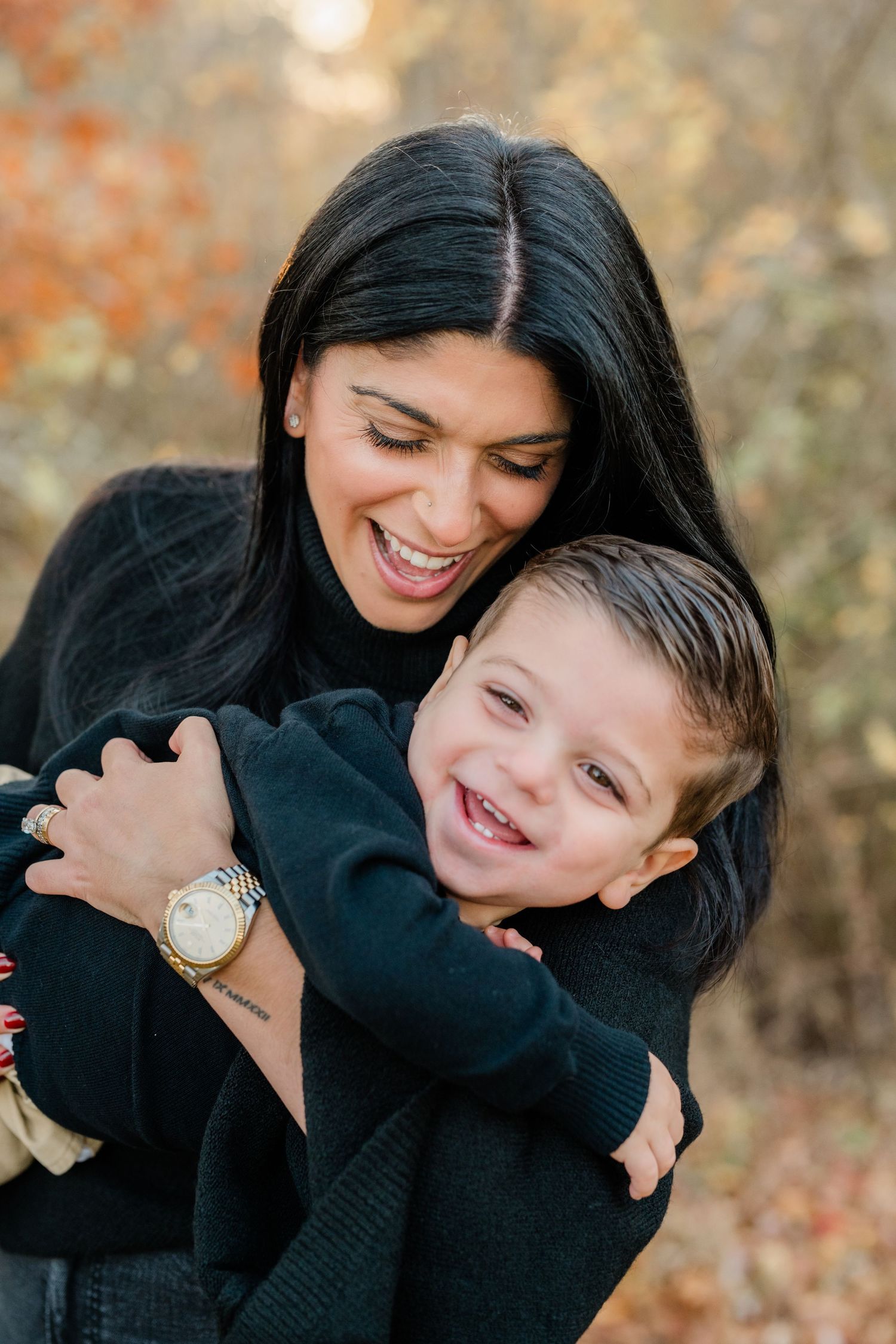 A laughing woman in black hugs a smiling young boy outdoors among autumn foliage.