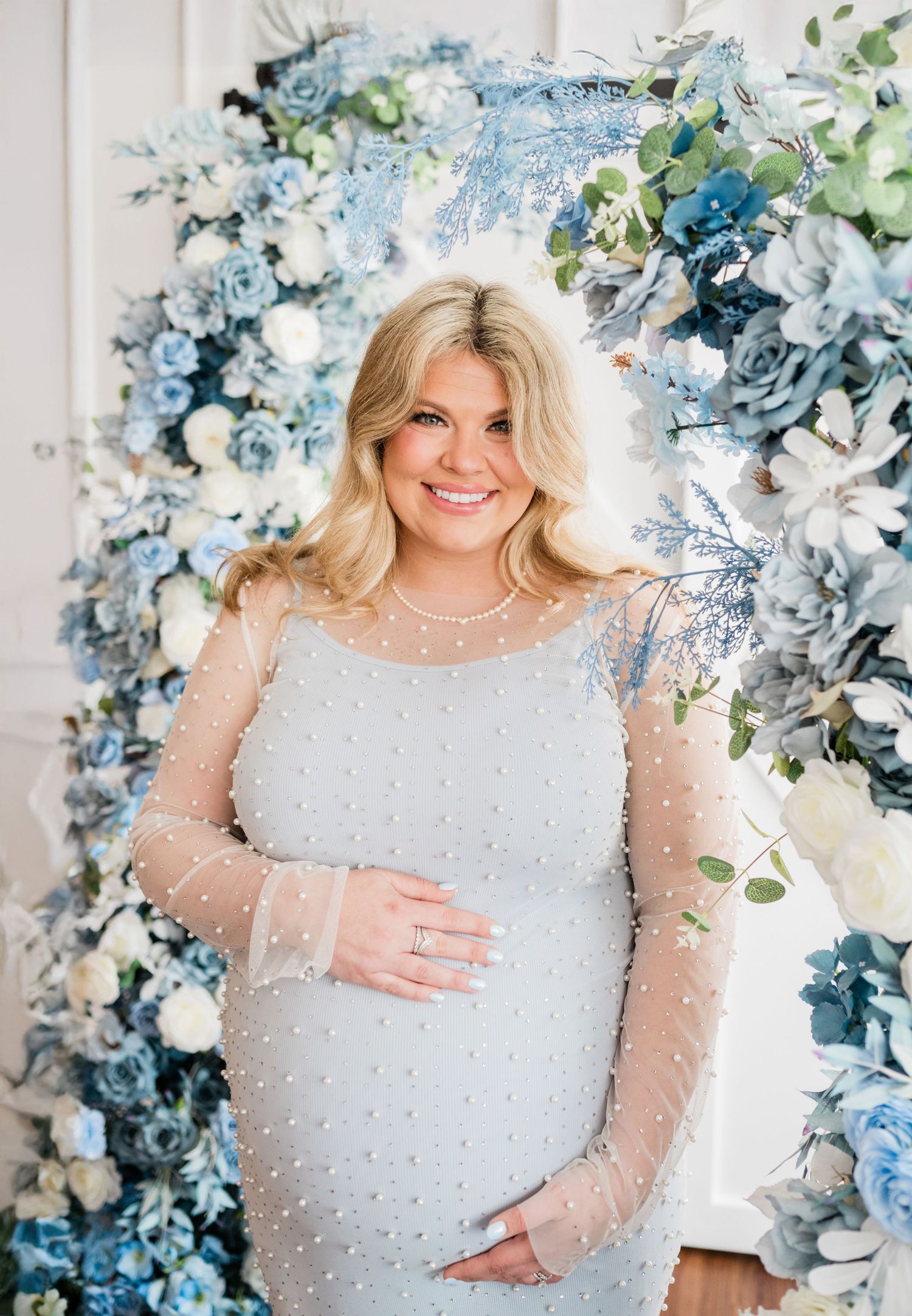 Maternity photos with woman in pearl-studded white dress against blue floral backdrop.
