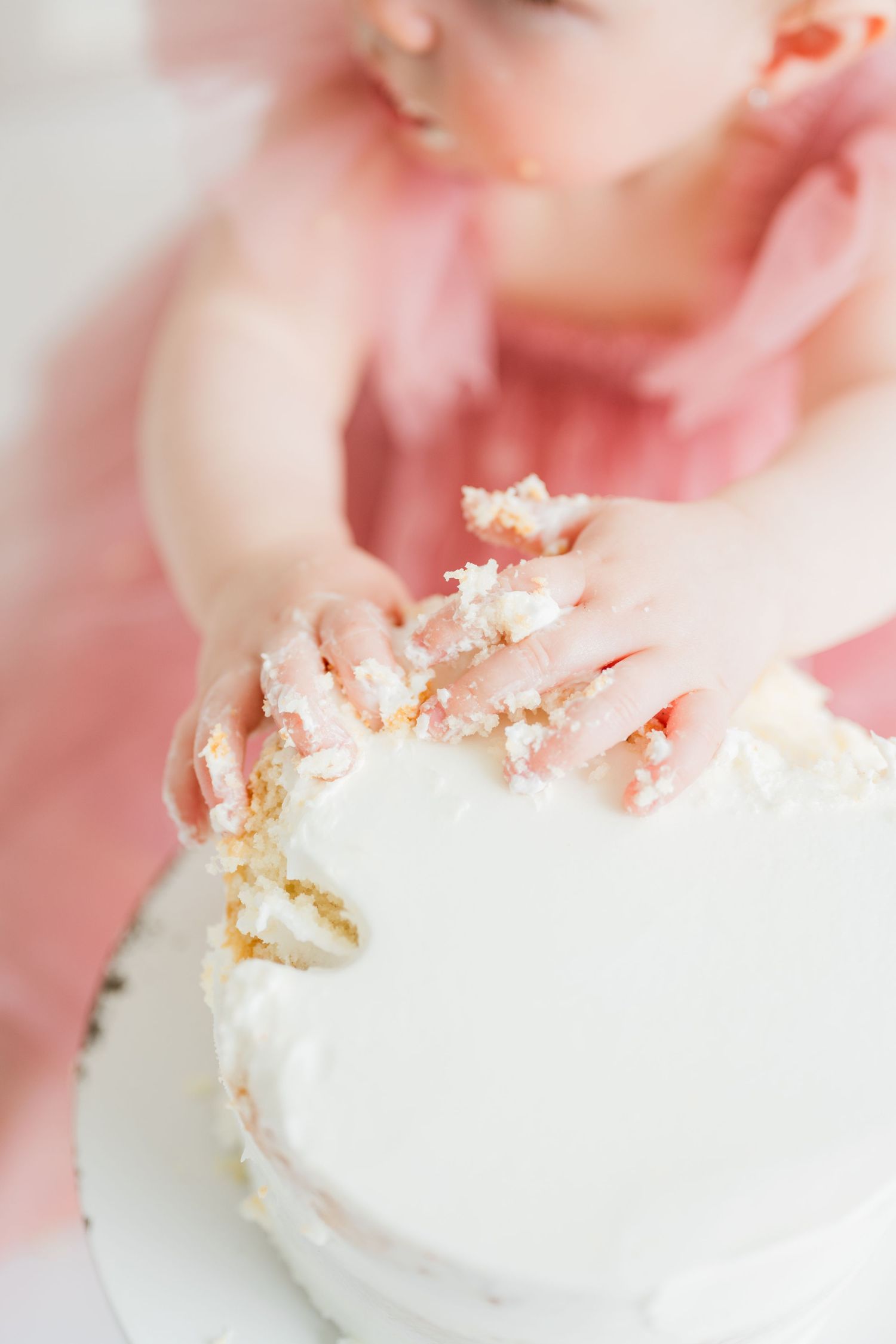 Baby in pink dress smashing white birthday cake with both hands during first birthday cake smash photoshoot.