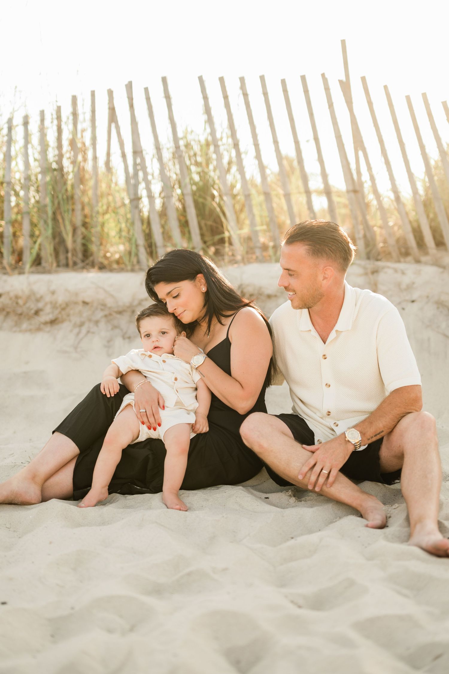 A young family sits on sandy beach dunes, mother holding baby while father smiles, with wooden fence backdrop.