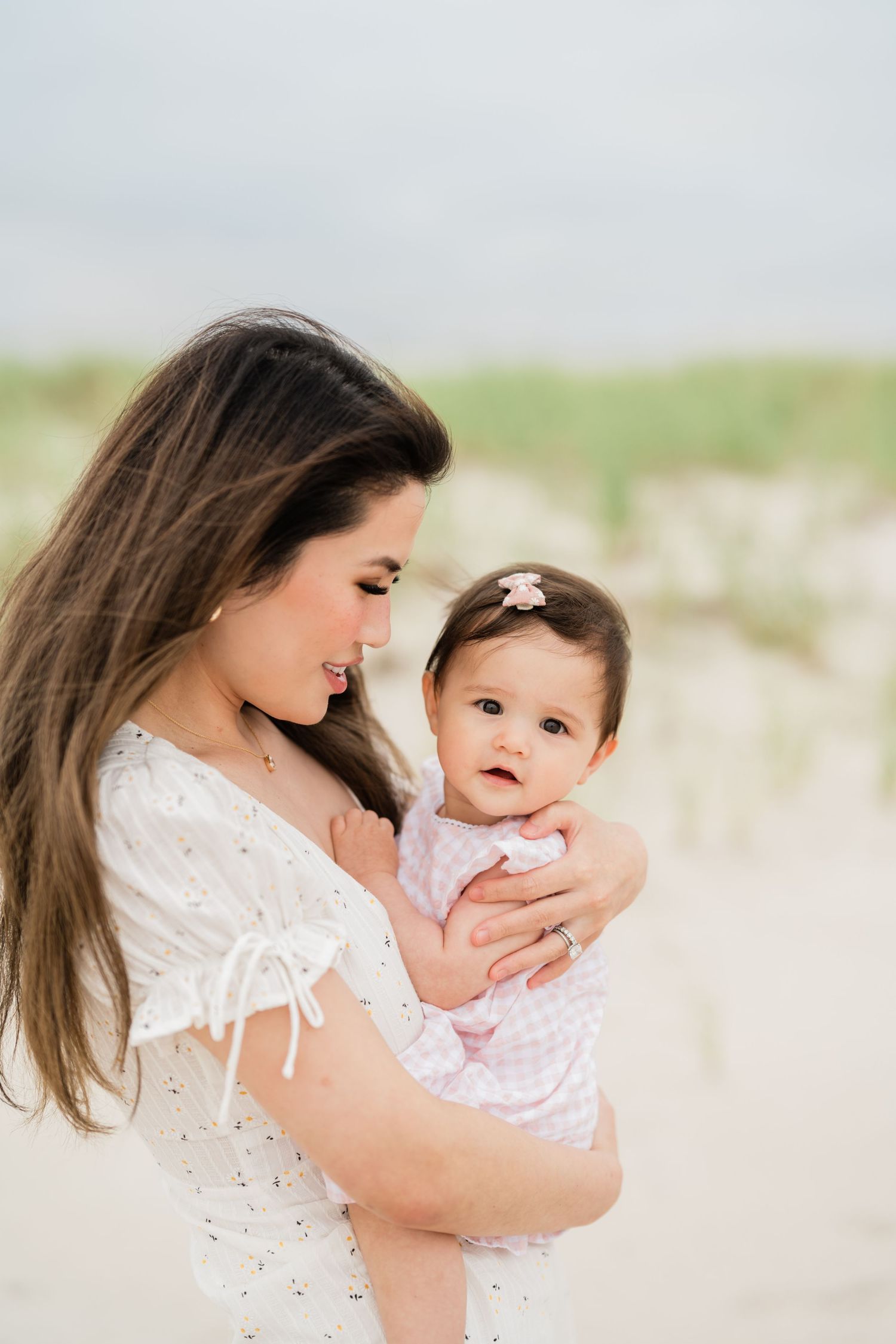 A mother in a white dress holds her baby girl in a pink outfit on a sandy beach.