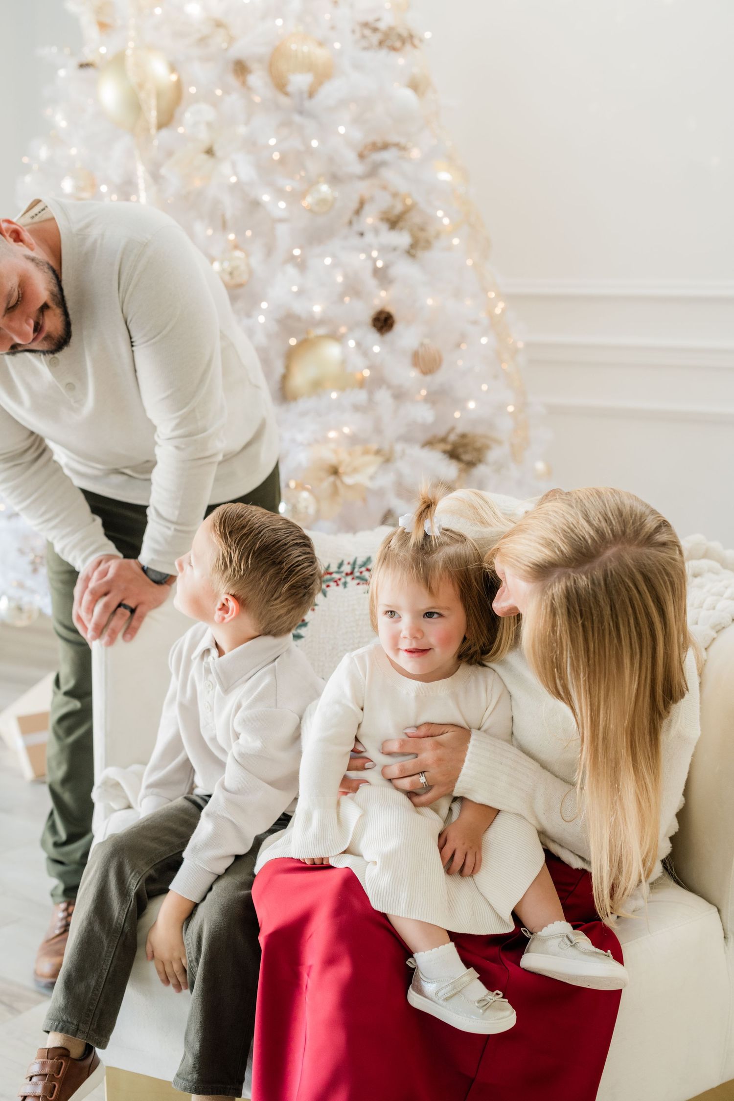 Family in white outfits celebrating Christmas near a decorated white and gold tree with two young children.