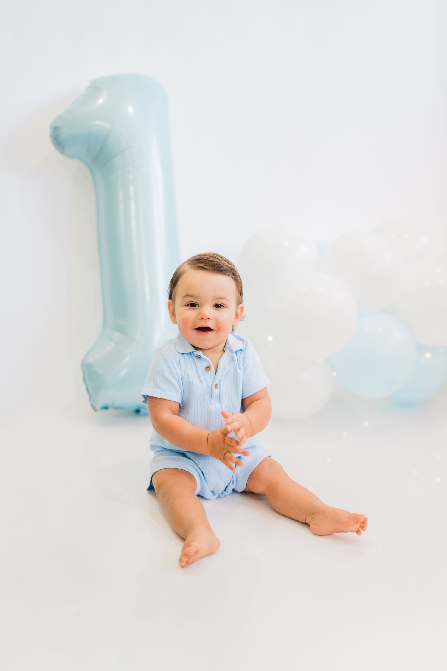 Smiling baby boy in blue romper sits on white floor beside large number 1 balloon celebrating his first birthday.