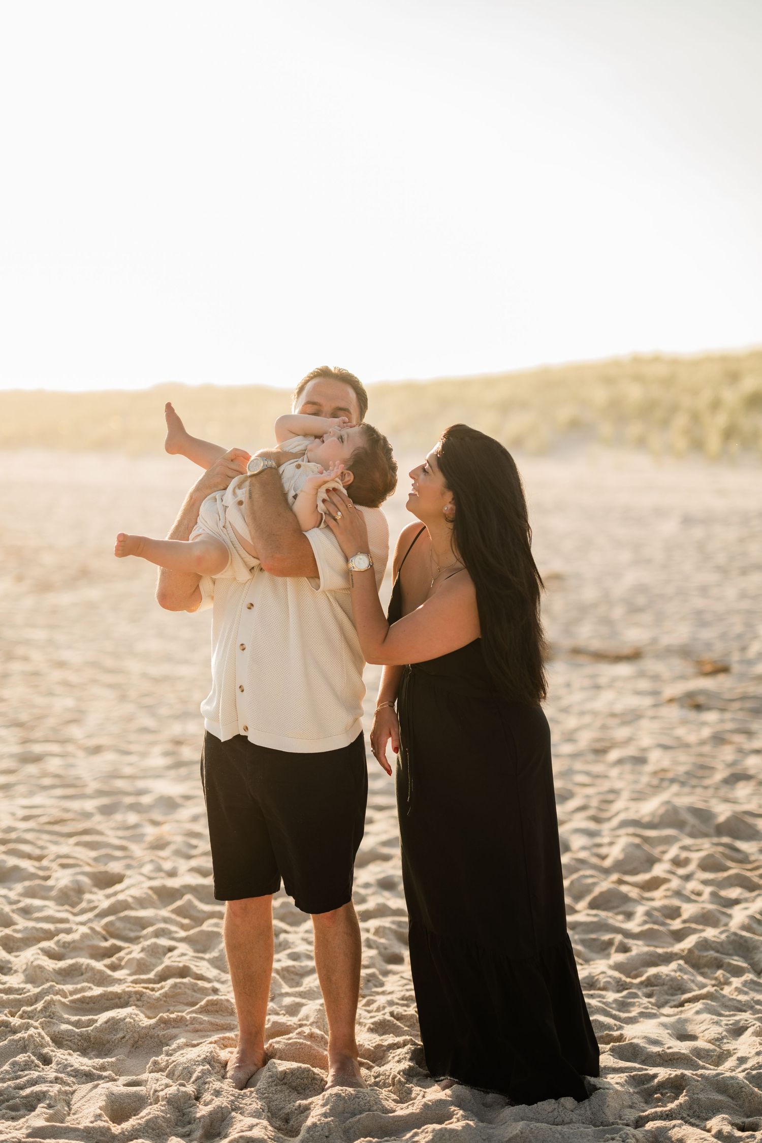 Family enjoying a golden sunset beach session, father lifting baby while mother in black dress looks on with joy.