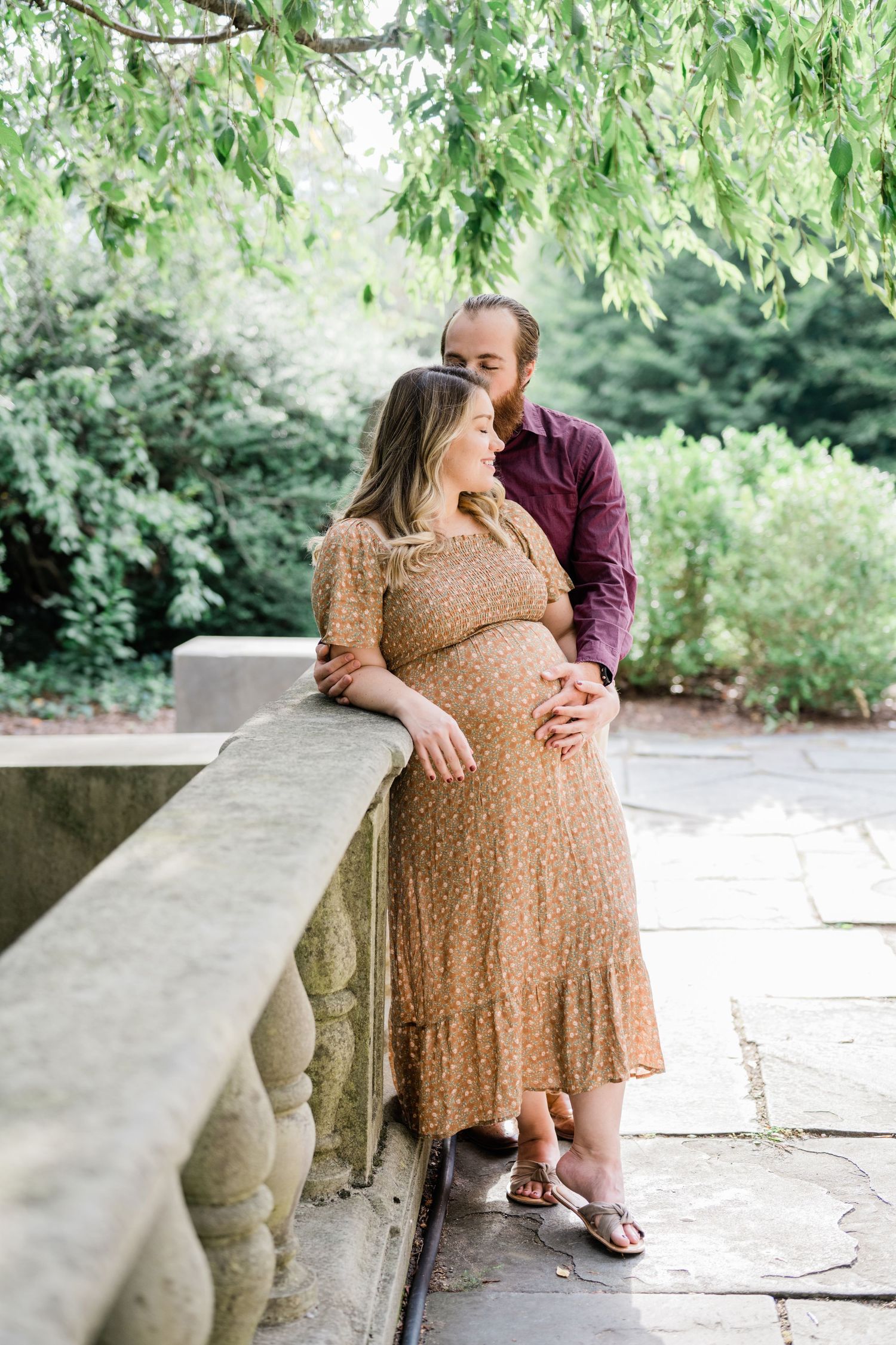 Romantic outdoor maternity photos on stone bench surrounded by lush greenery.