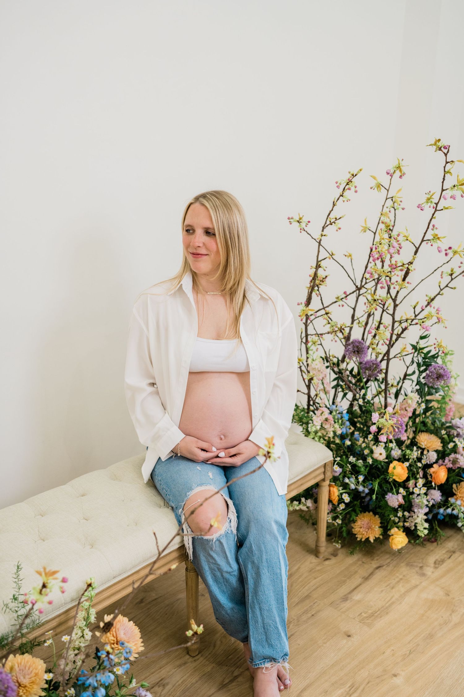 Maternity photoshoot featuring floral arrangements and casual styling against a white backdrop.