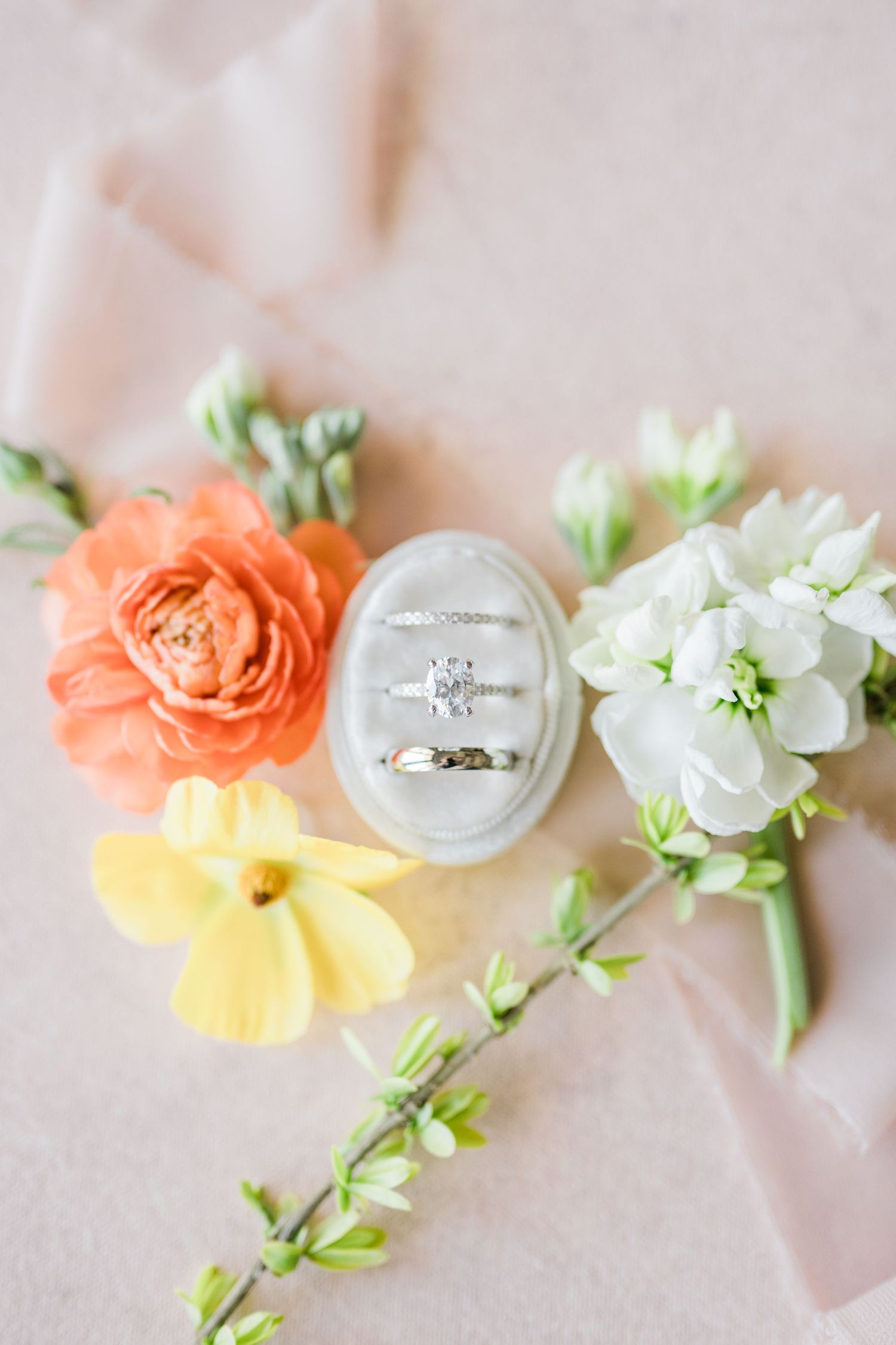 Wedding ring detail shot with coral, yellow and white flowers arranged on light pink background.