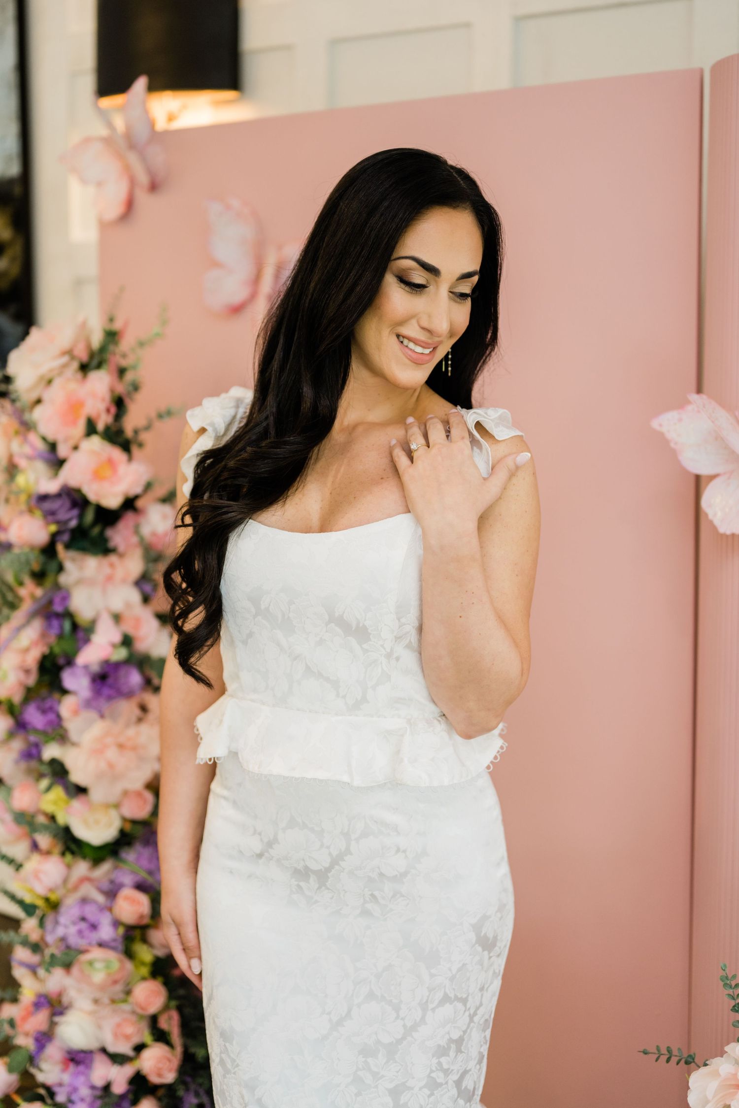 A bride in a white lace peplum dress poses against a pink backdrop with floral decorations.