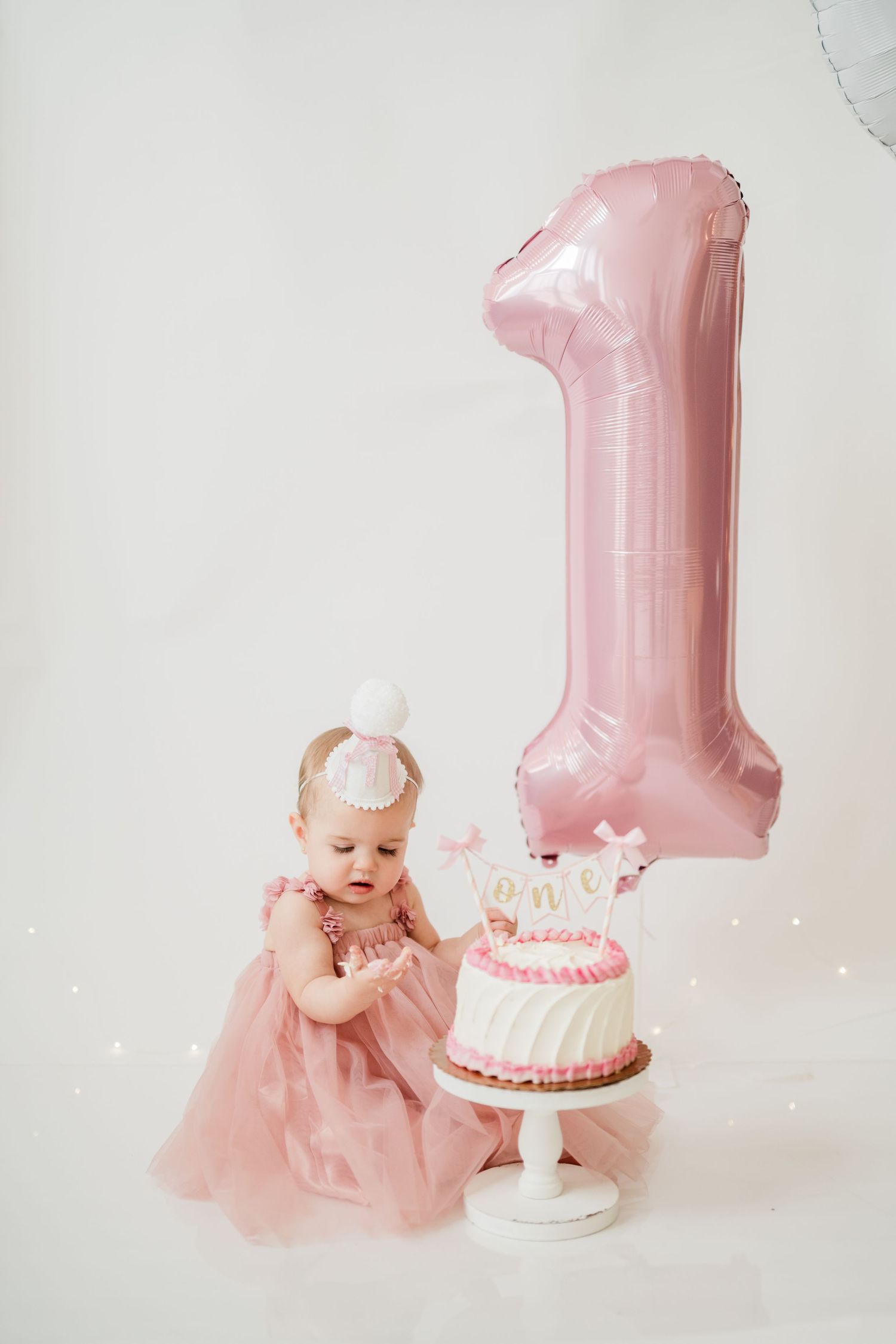 Baby girl in pink dress celebrates first birthday with cake and number one balloon.