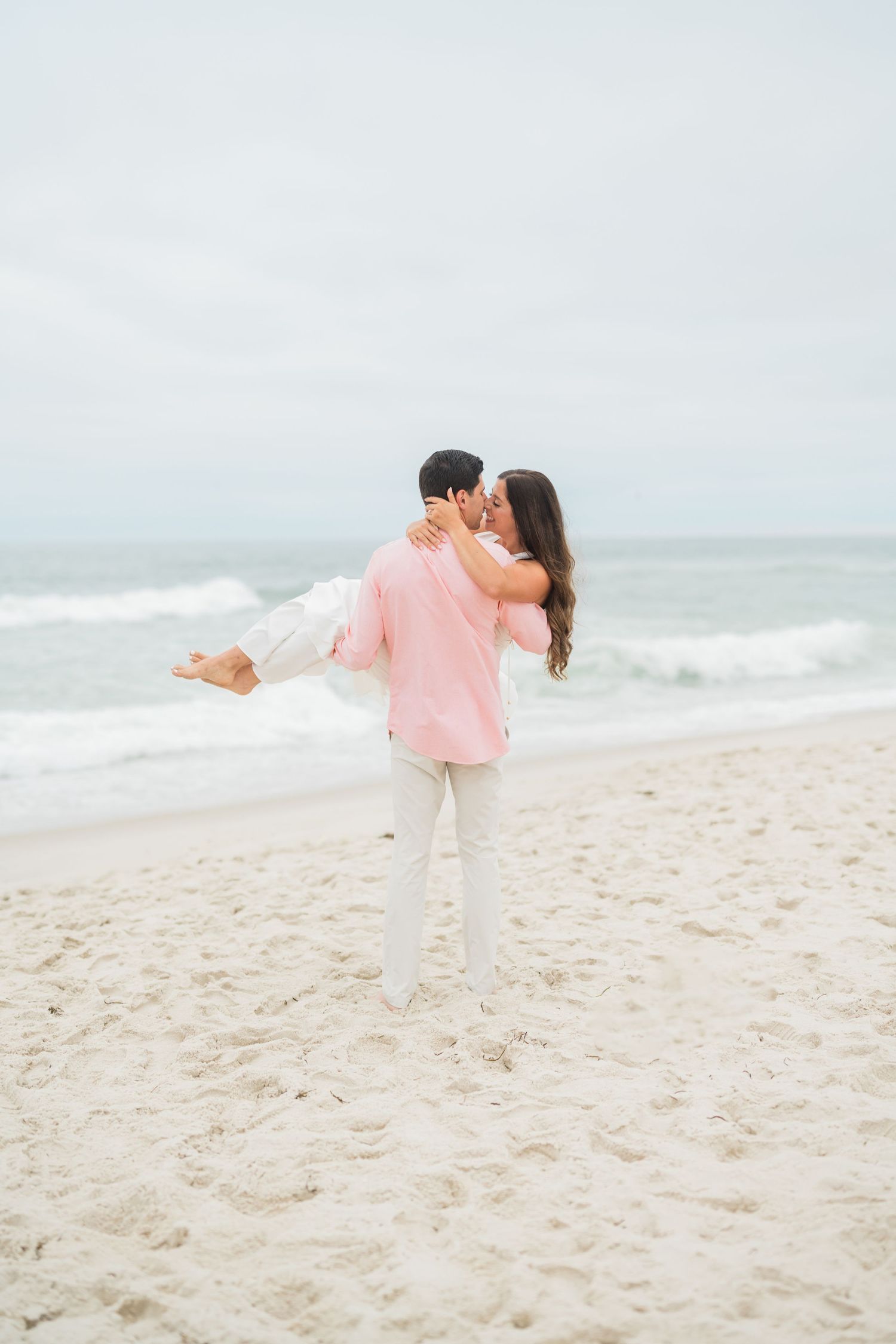 A man in a pink shirt carries a woman in white on a sandy beach, sharing a romantic kiss with ocean waves behind them.