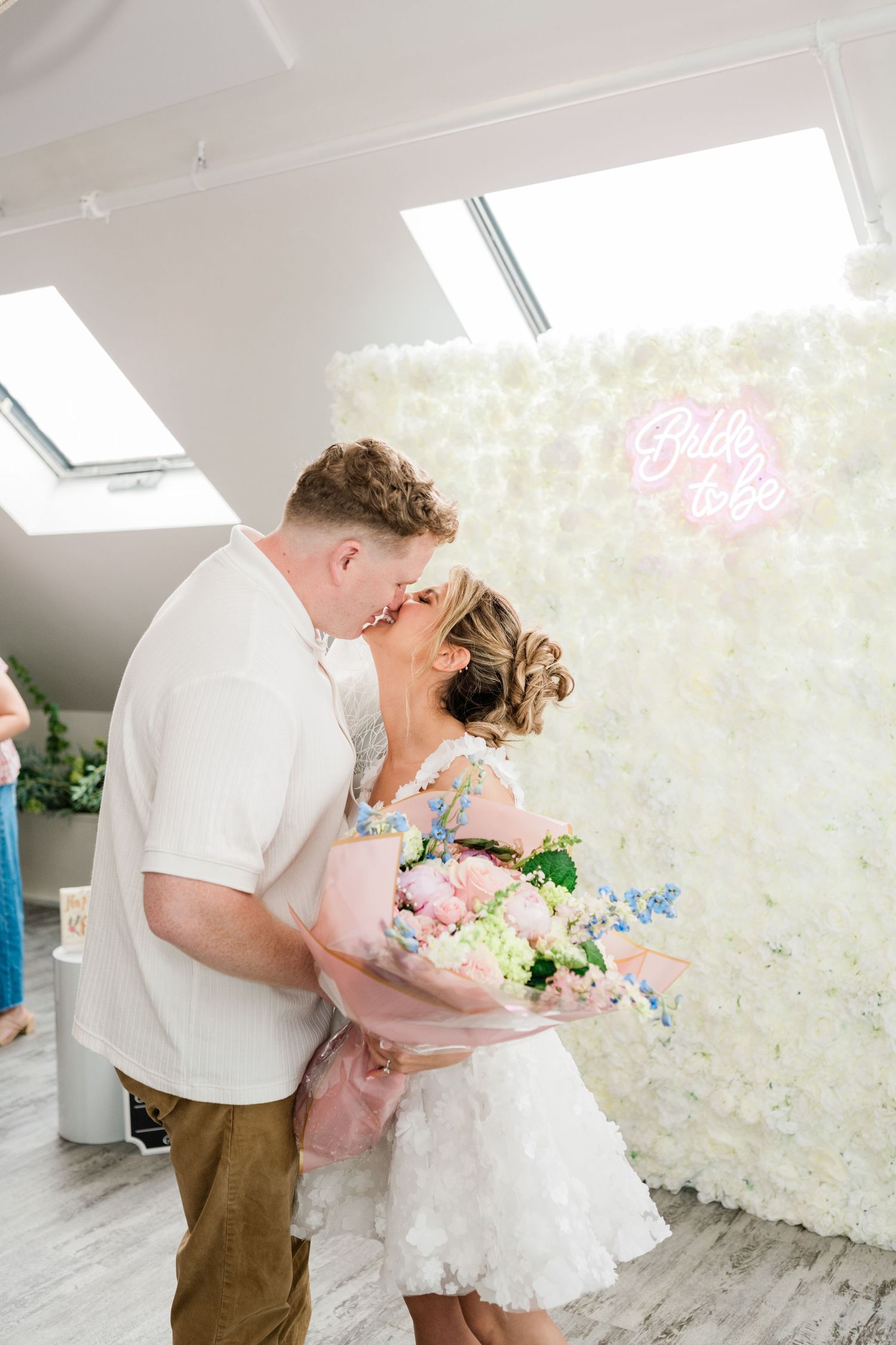 A romantic photoshoot in a bright room with white floral wall decorations and a pink neon sign.