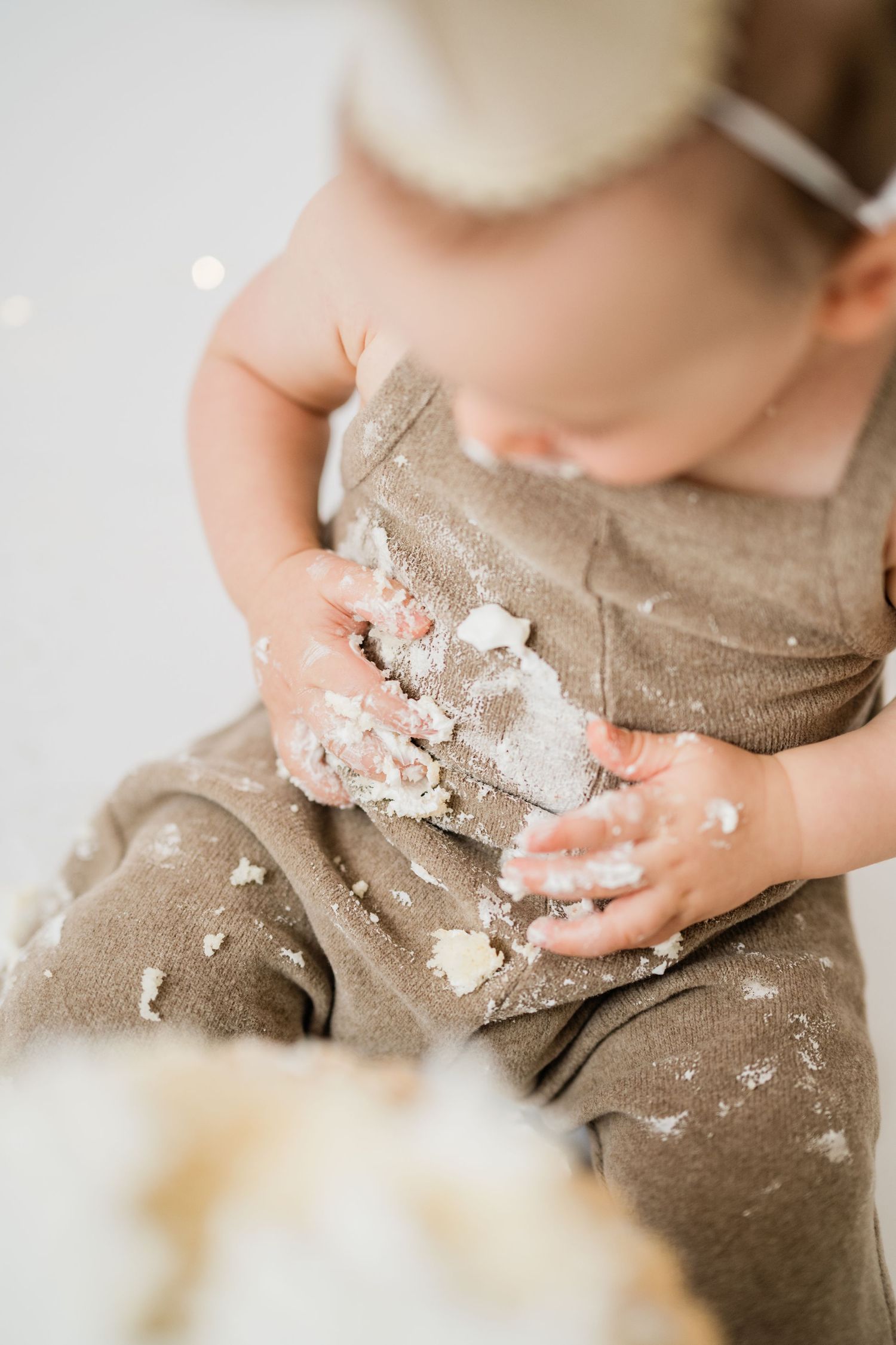 Baby in tan romper smashing first birthday cake with both hands, covered in white frosting.
