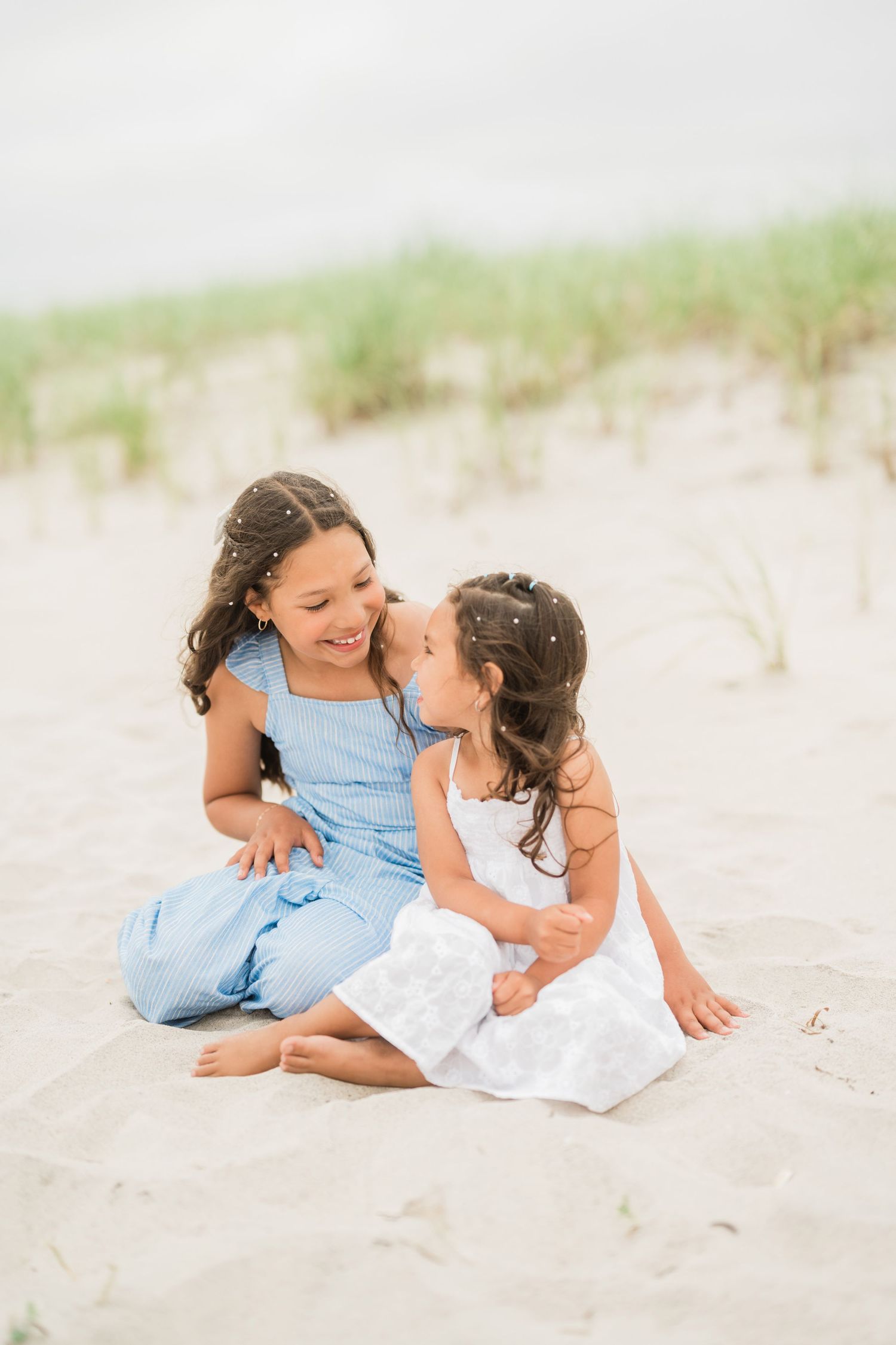 Two young sisters in blue and white dresses sitting together on sandy beach dunes, smiling and sharing a sweet moment.
