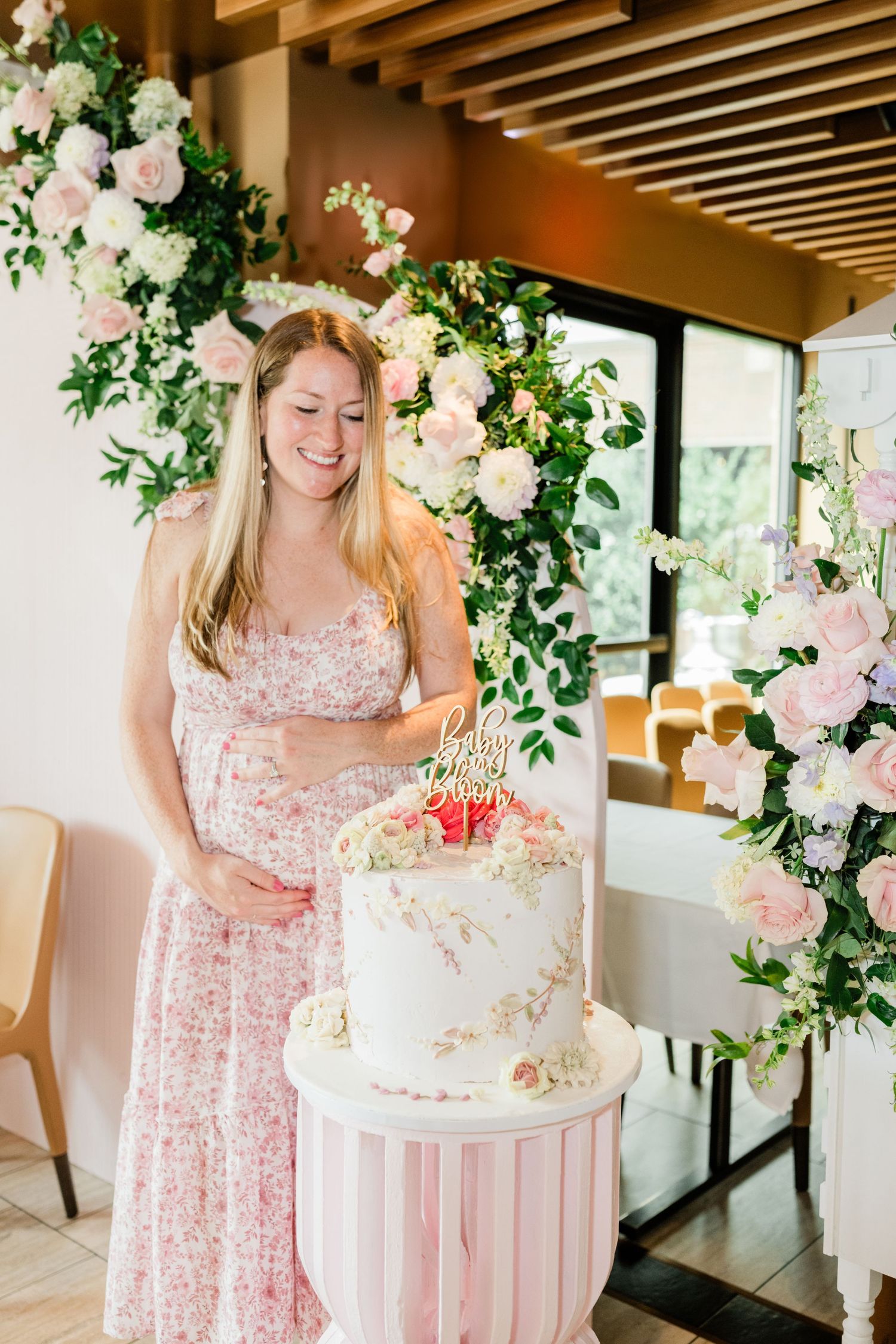 Baby shower celebration with pink floral decor and white tiered cake on pink striped stand.