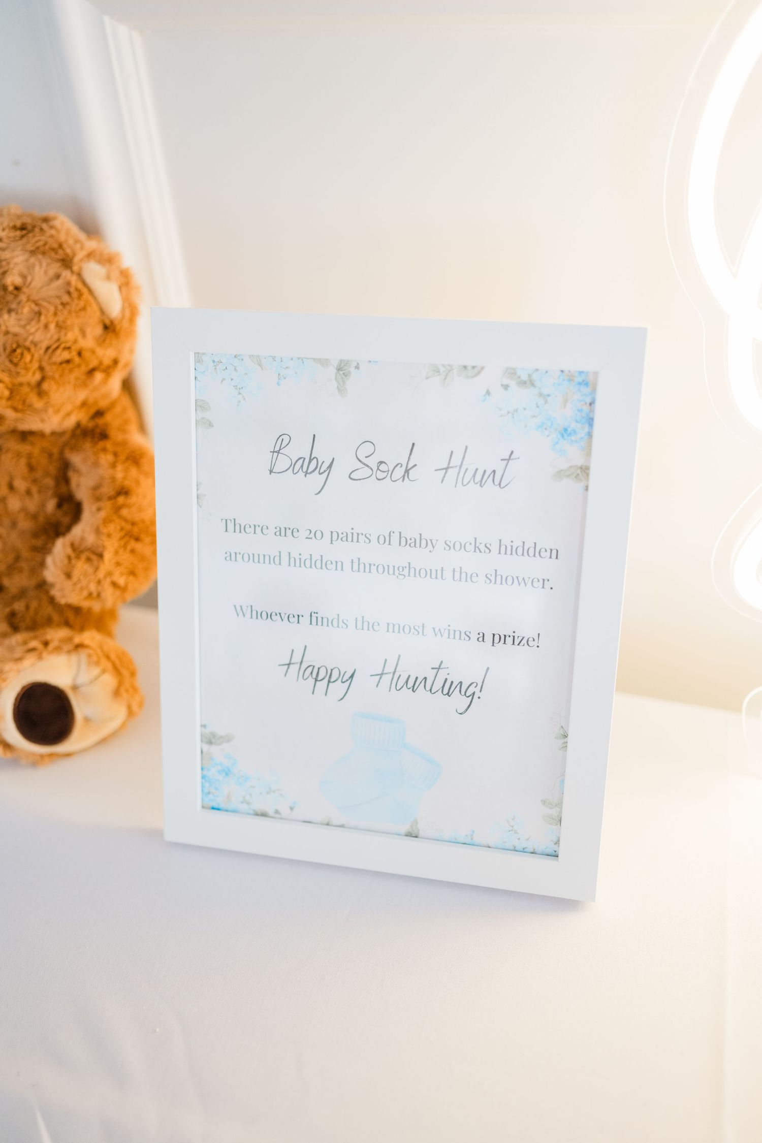 Baby sock hunt instruction card with blue floral border next to plush teddy bear.