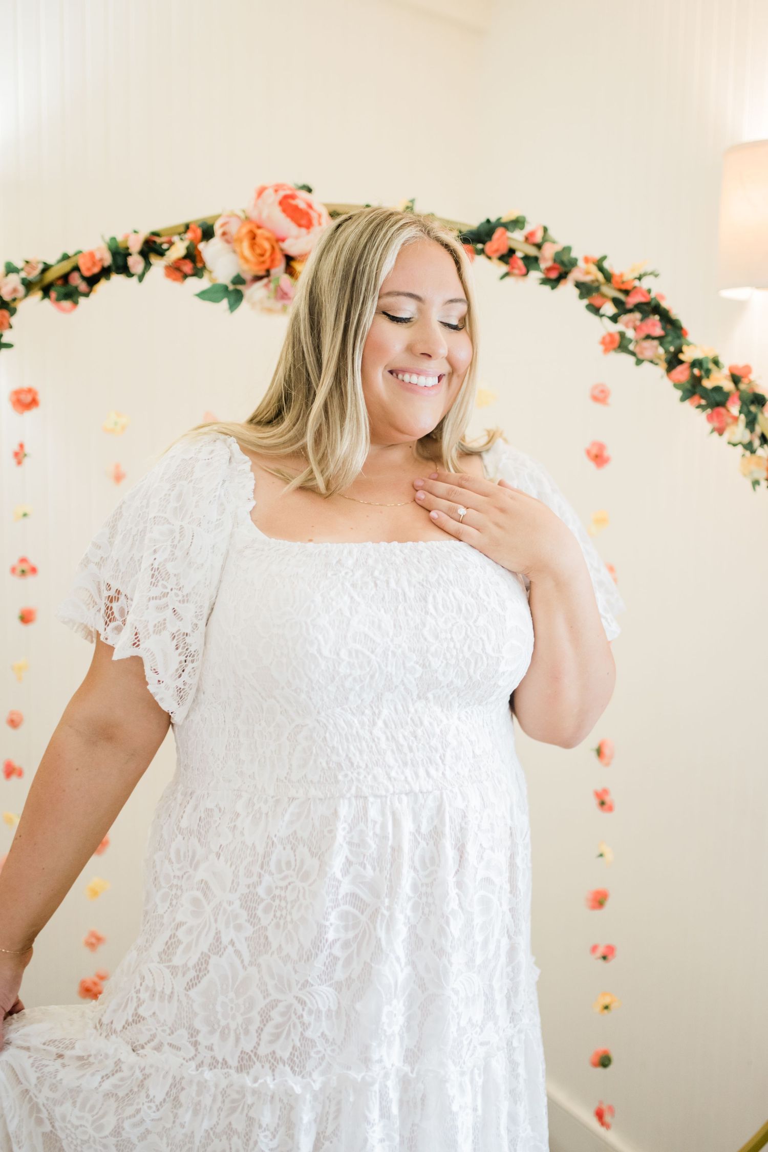 Woman in white lace dress smiling under floral arch with peach and coral flowers.