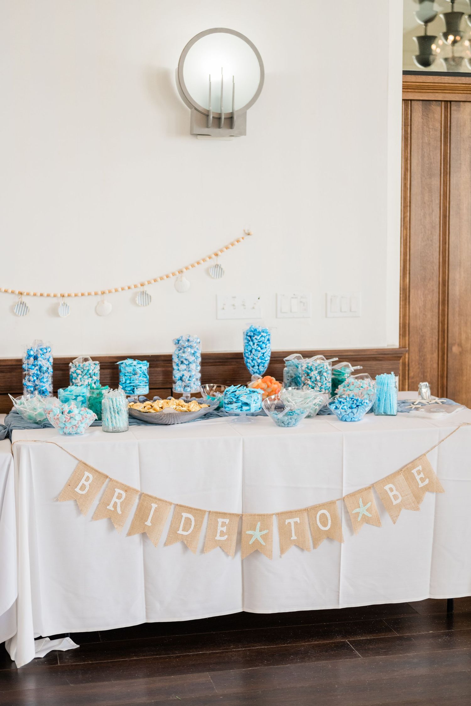 Elegant beach-themed party table setup with blue decor and burlap banner against white walls.